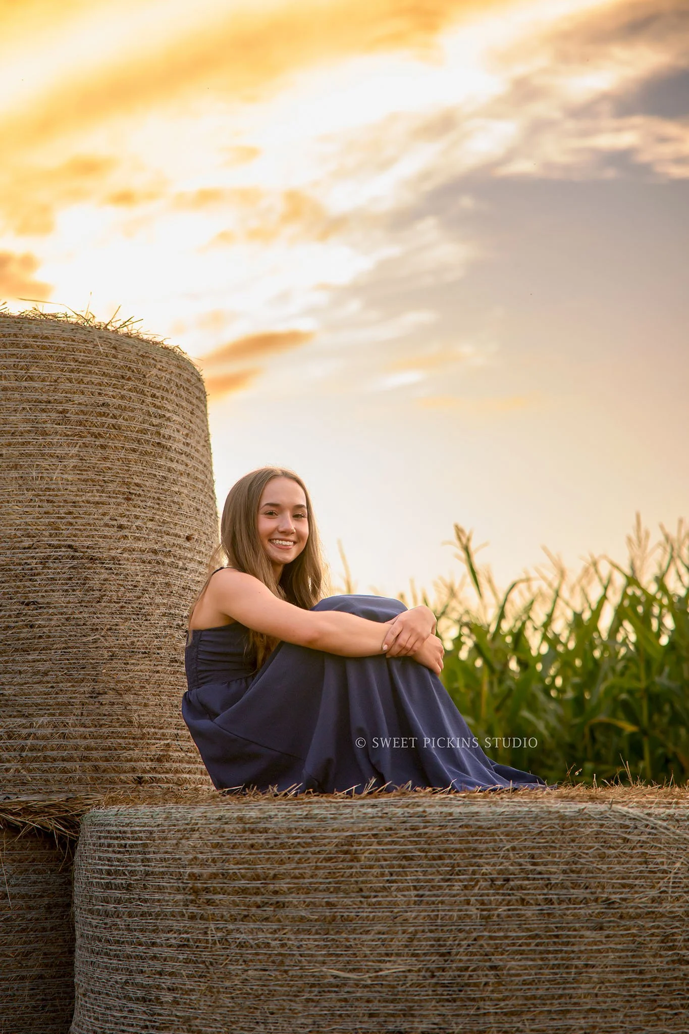 Natalie’s Lowell, Indiana Farm Senior Photography posing on hay bales by corn in blue dress at sunset  by Sweet Pickins Studio senior photographer Amanda Wilson ©