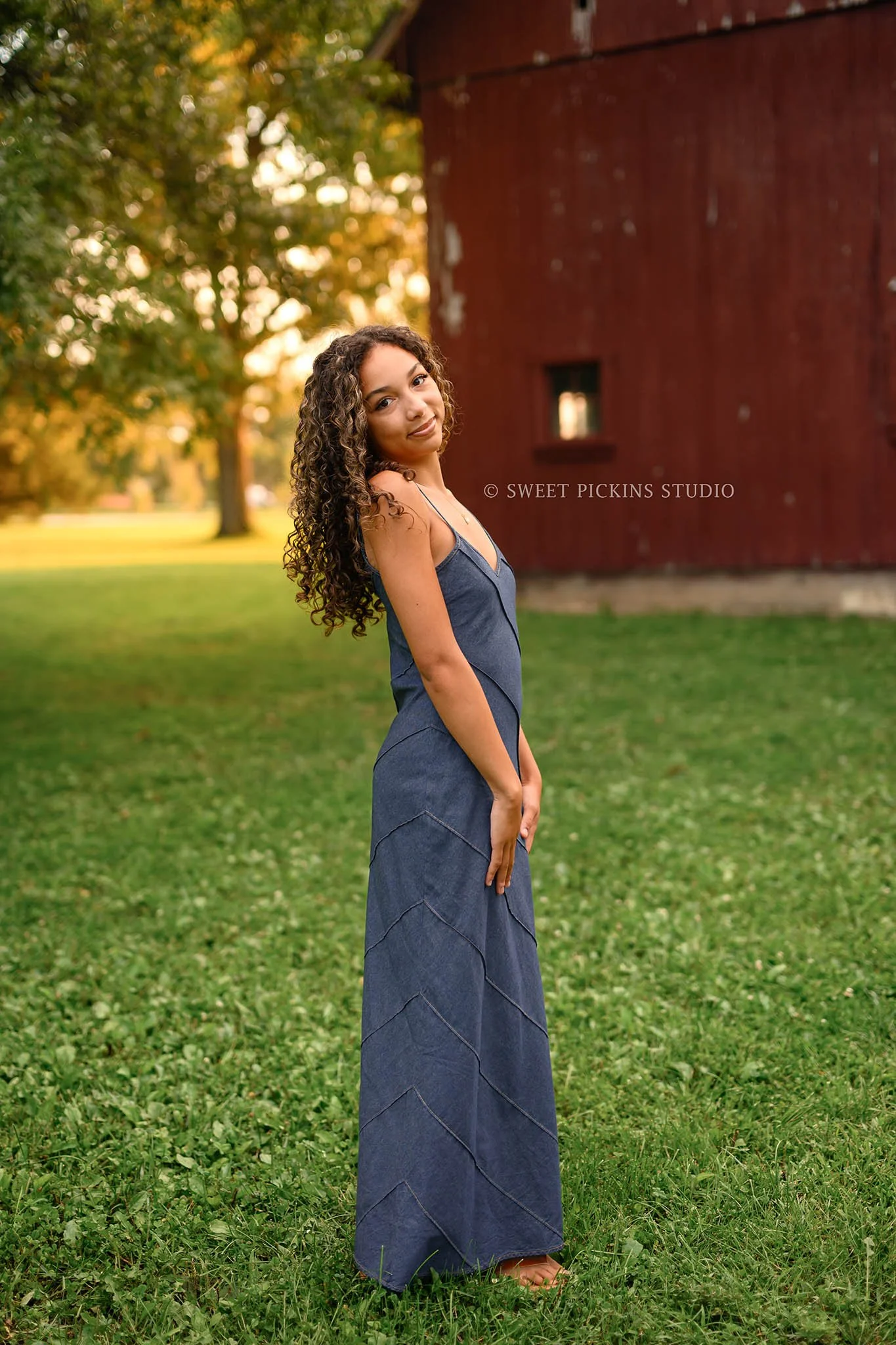 Madeline | Senior Photography for Class of 2027 senior girl with curly hair wearing denim jean dress near barn by Peru Indiana Senior  photographer Amanda Wilson of Sweet Pickins Studio ©