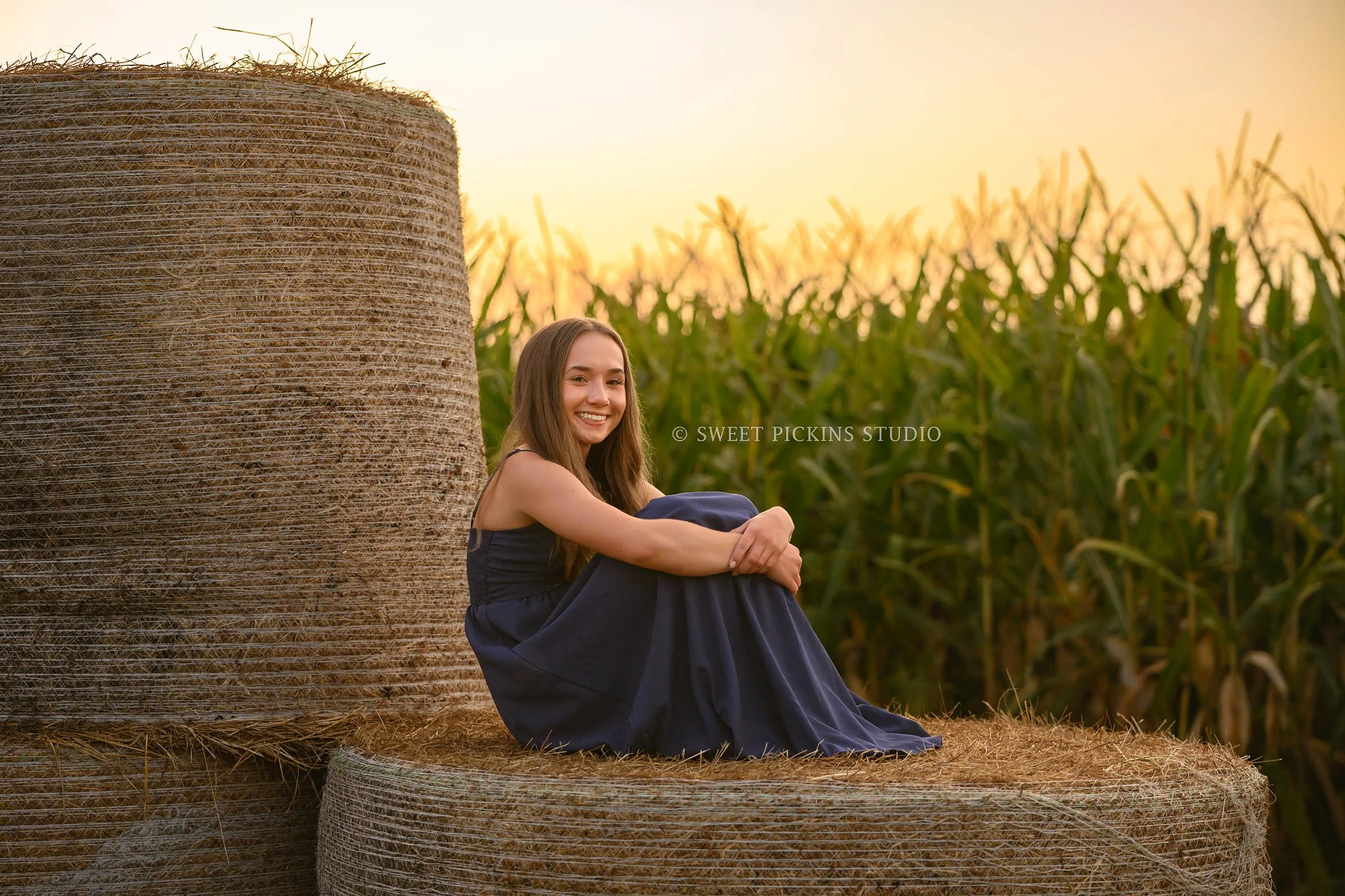 Natalie’s Lowell, Indiana Farm Senior Photography posing on hay bales by corn in blue dress at sunset  by Sweet Pickins Studio senior photographer Amanda Wilson ©