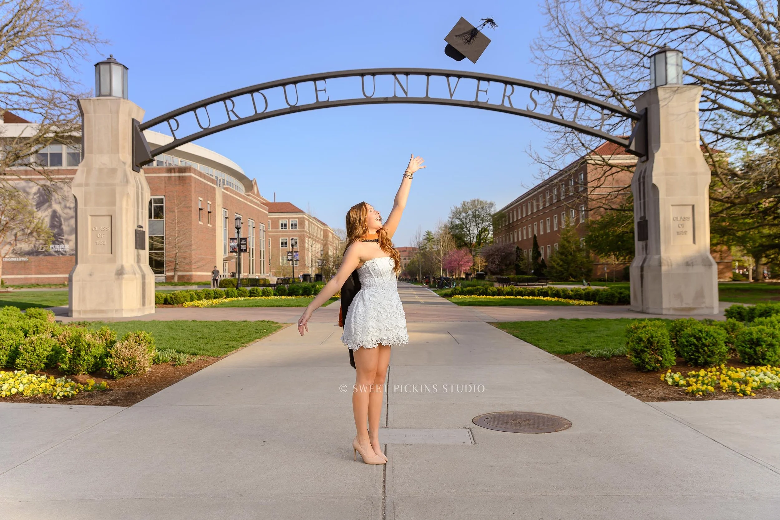 Lauren’s Purdue University Masters Graduation Photography in West Lafayette, Indiana by Sweet Pickins Studio photographer Amanda Wilson for graduate tossing cap in white dress at Engineering arch wearing stole © 2026