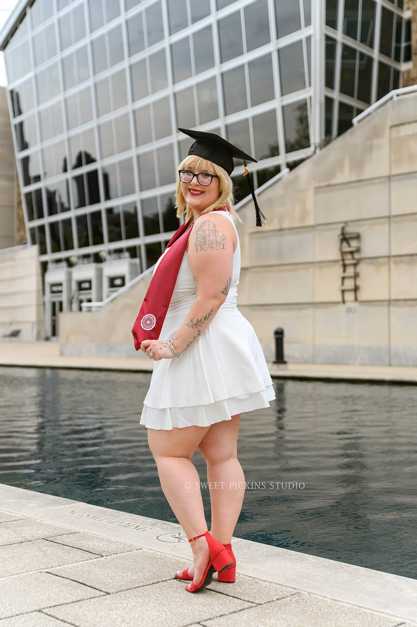 I.U. Indy Graduation Photography in Downtown Indianapolis, Indiana for class of 2026 spring grad wearing red high heels by canal water by Sweet Pickins Studio senior photographer Amanda Wilson ©