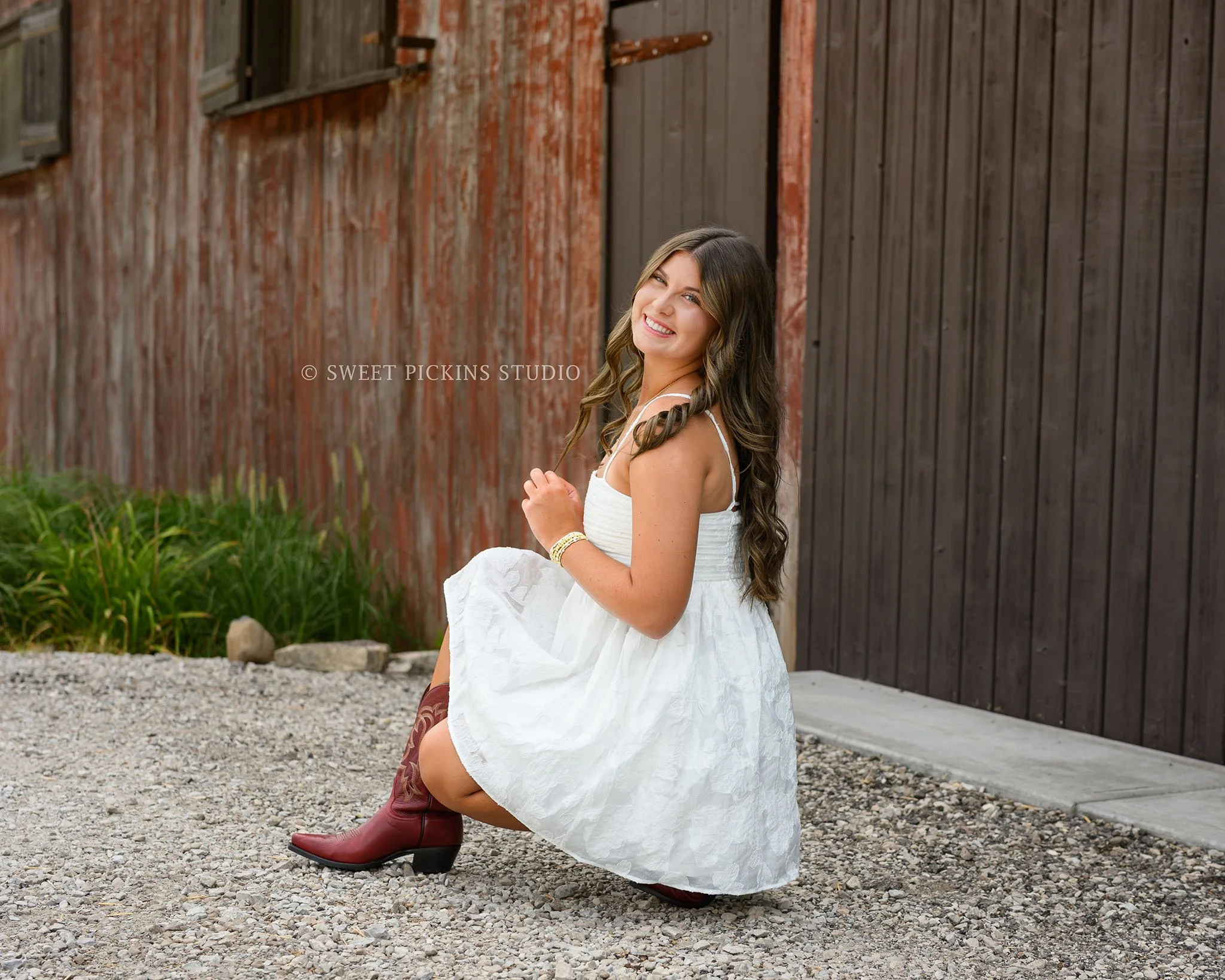 Zionsville, Indiana Country Senior Portrait Photography at Traders Point Creamery with class of 2027 senior girl posing in white babydoll dress and red western boots by red barn by Sweet Pickins Studio senior photographer Amanda Wilson ©