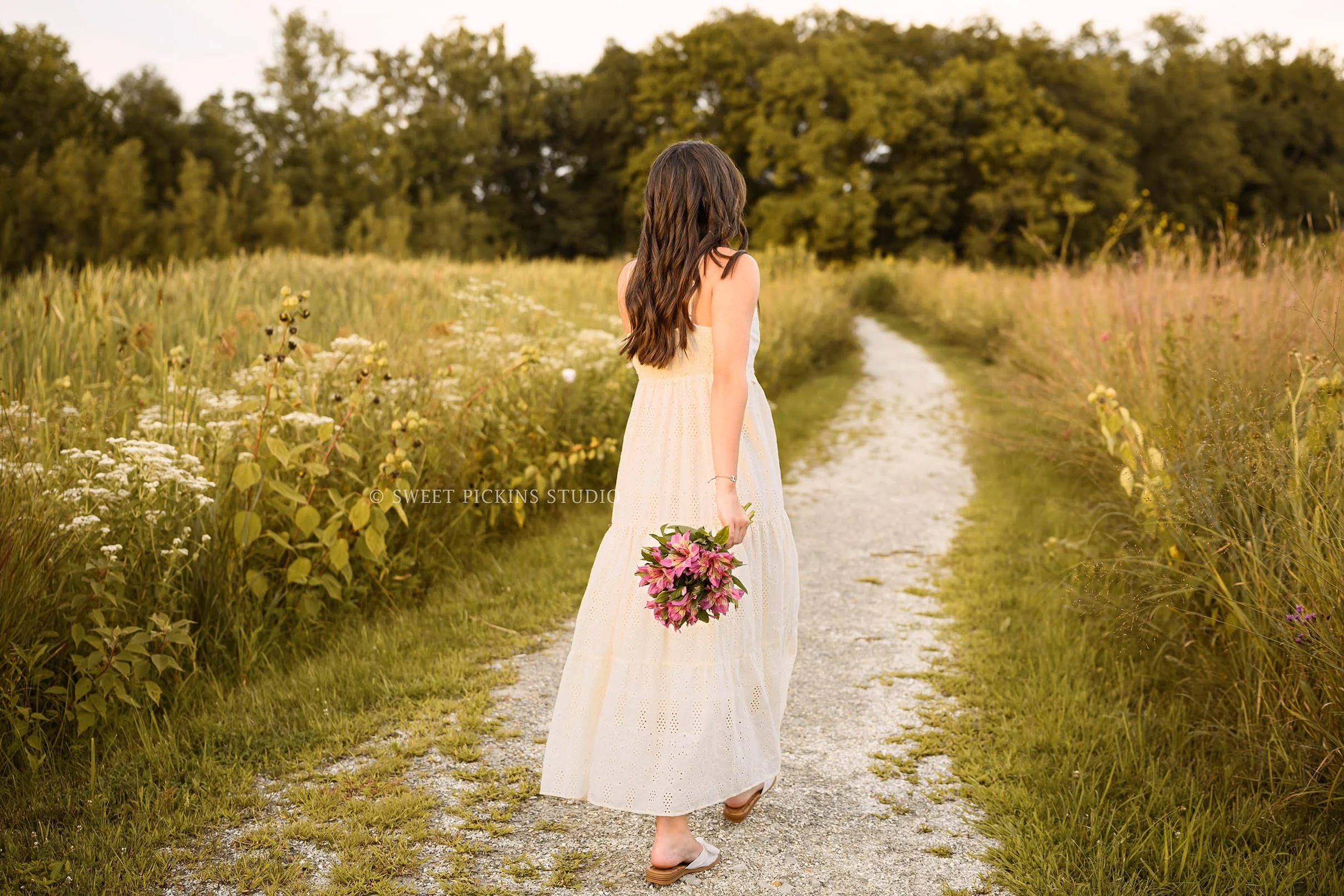 Sophie’s Carmel, Indiana Senior Photography by Sweet Pickins Studio senior photographer Amanda Wilson for class of 2027 Senior girl walking in flower field in cream maxi dress at sunset ©