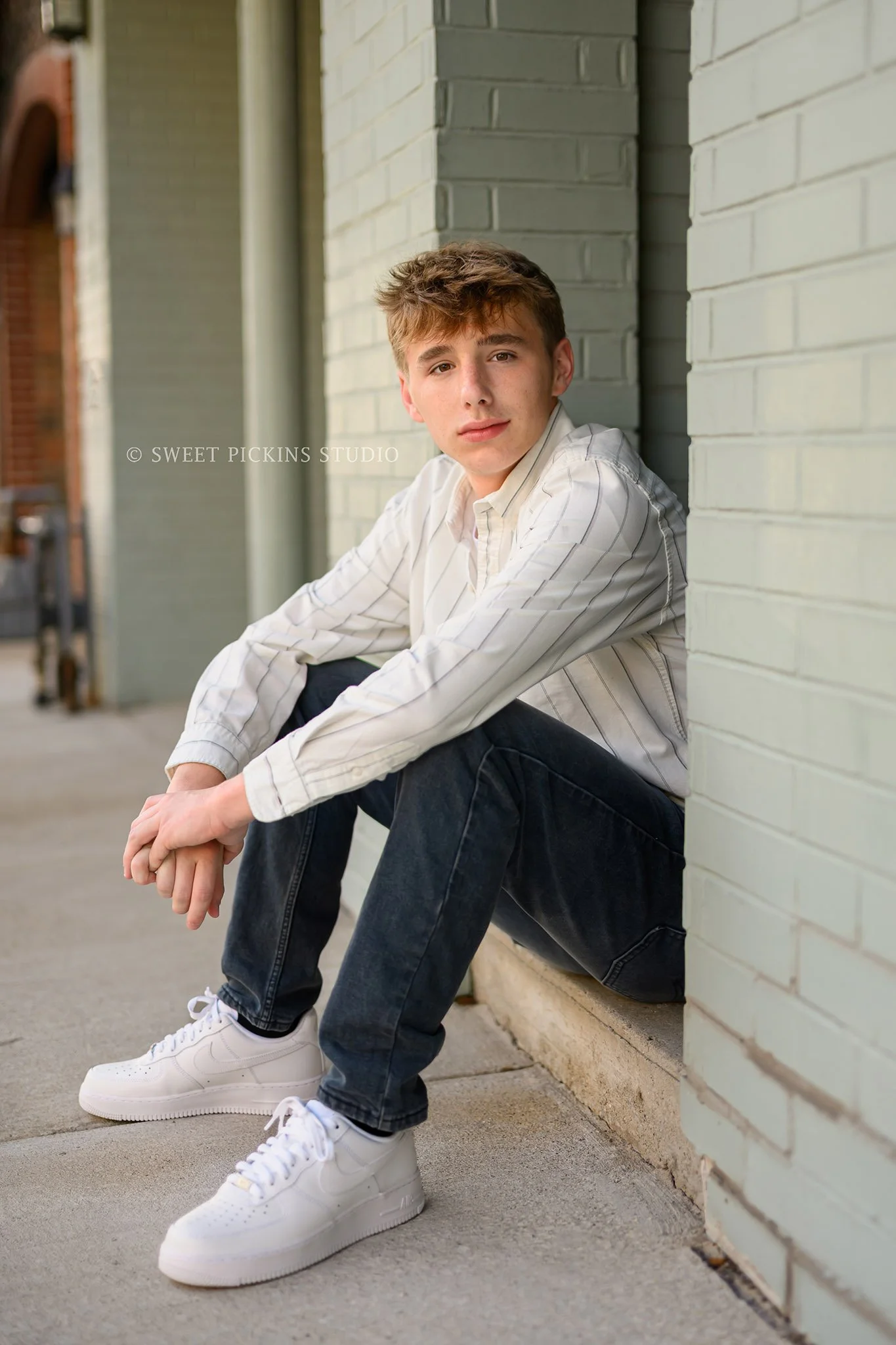 Huntington, Indiana Senior Guy Portraits for Huntington North Class of 2027 Senior boy BOSTAN sitting on  steps in denim jean button down shirt and white Nikes in urban downtown by Sweet Pickins Studio senior photographer Amanda Wilson ©