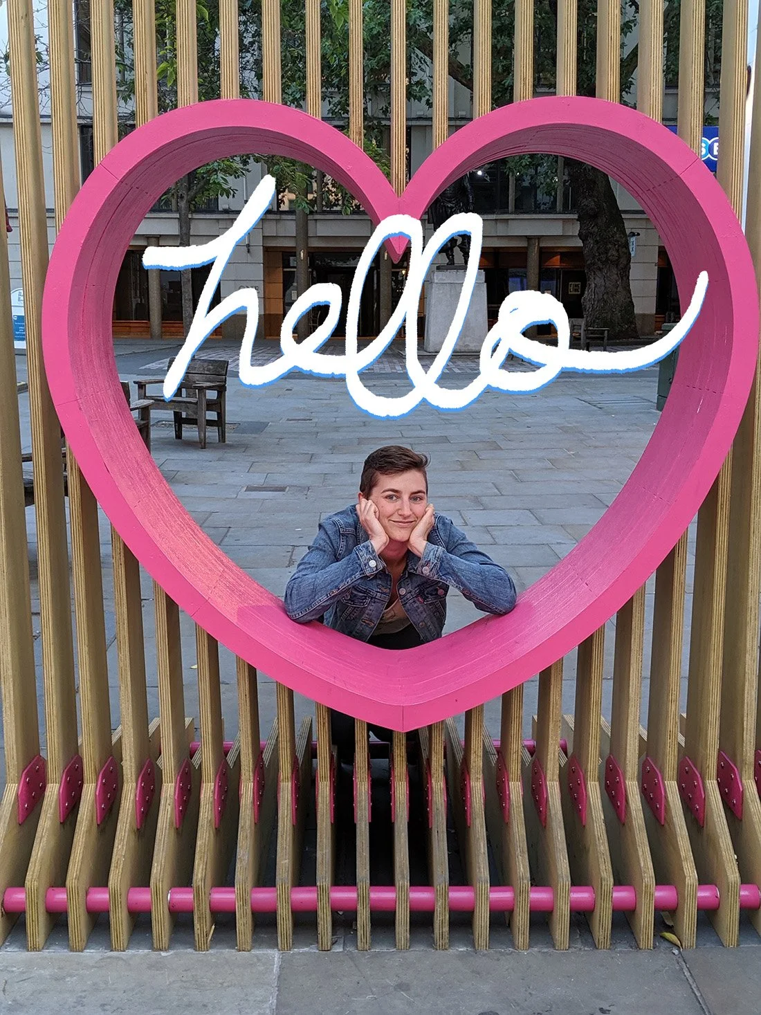 the artist, a short haired white woman in a denim jacket, leans on her elbows through a large wooden pink heart. hand lettering above her reads Hello