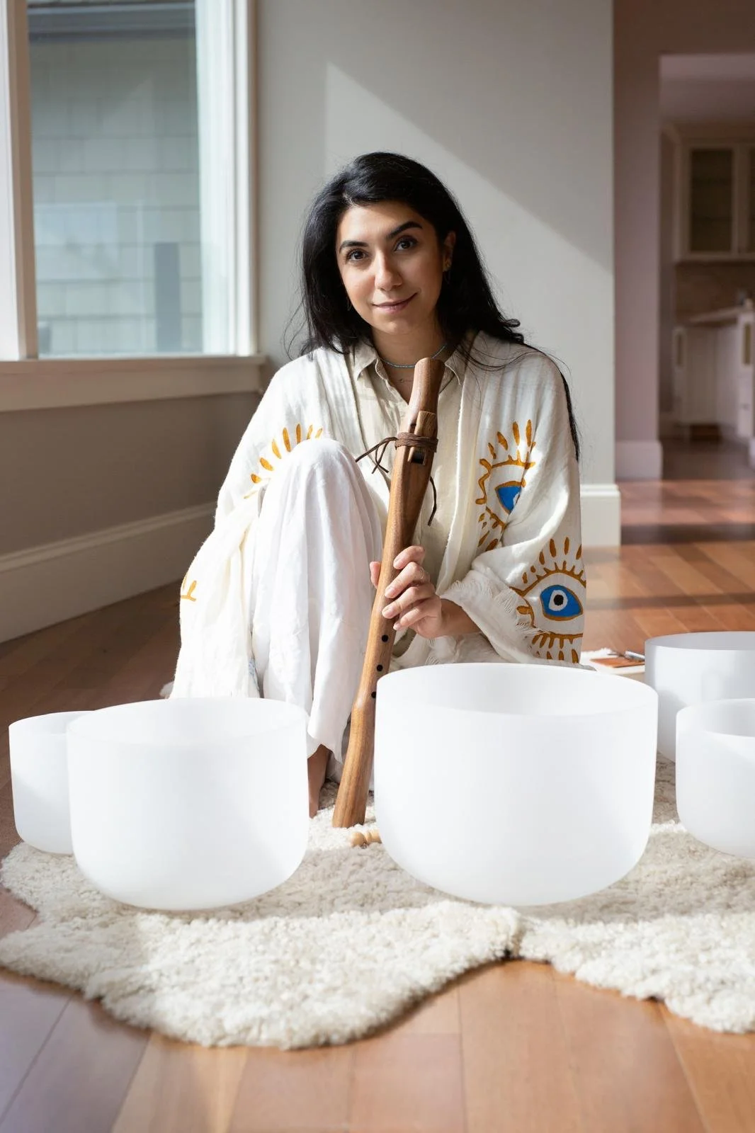 Woman seated on a rug with a wooden flute and crystal singing bowls.