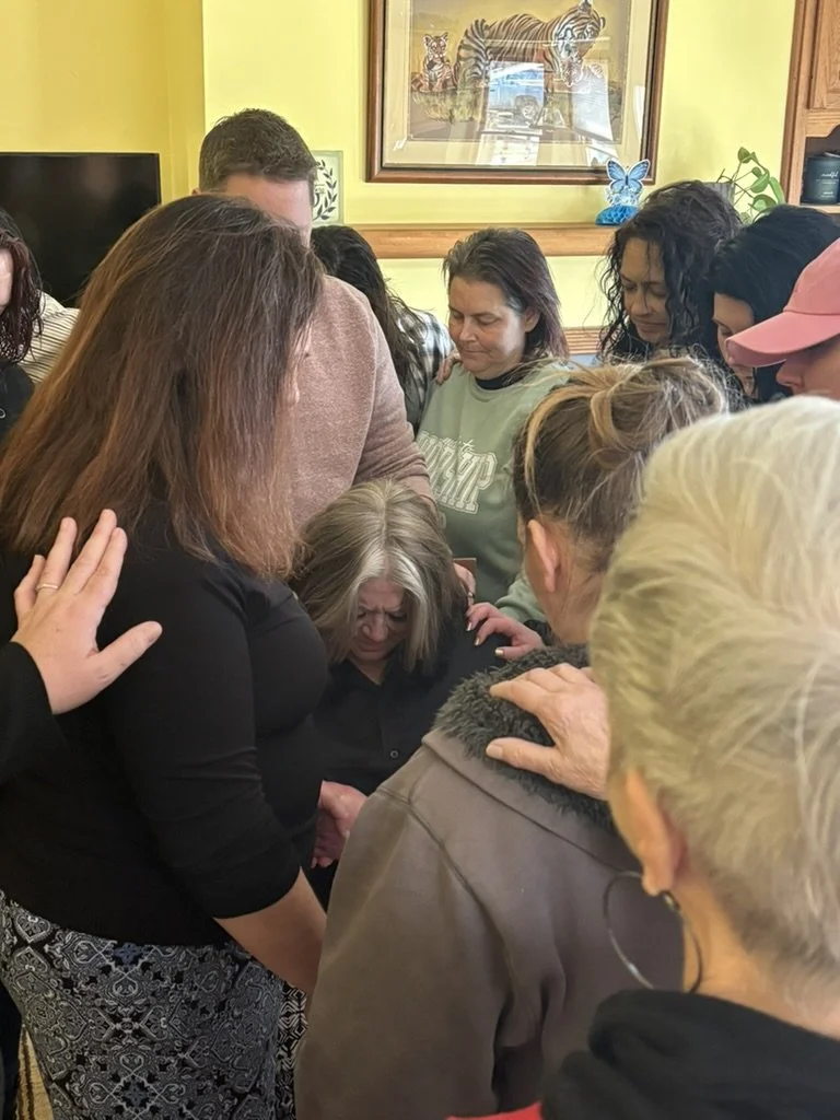 A group of women and one man gathered closely together, appearing to be in prayer or deep reflection, with a woman in the center looking down with a somber expression.