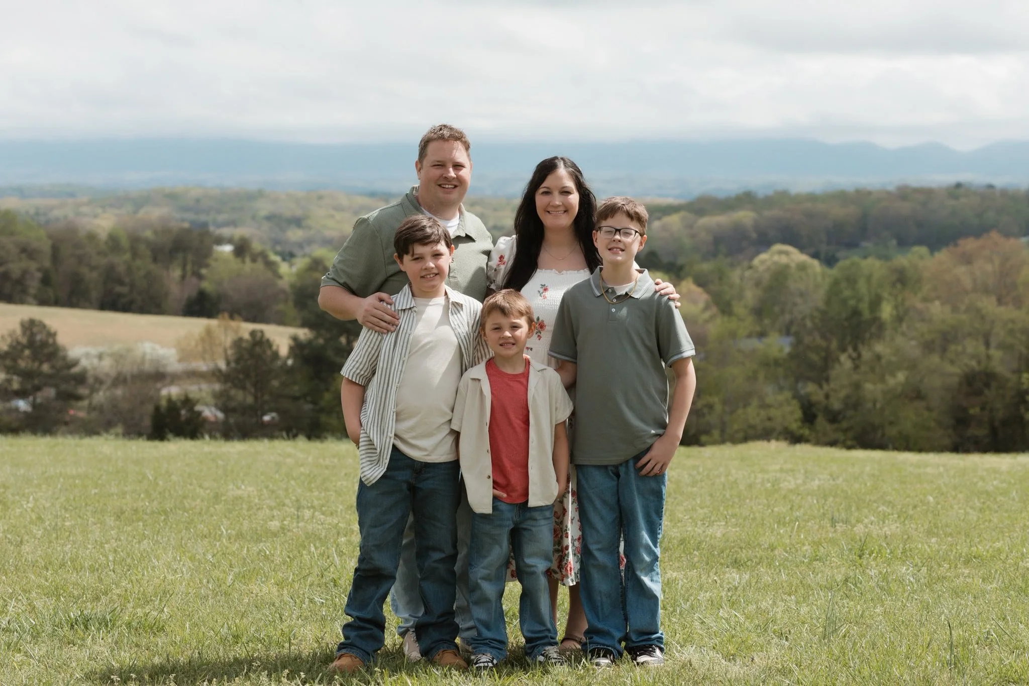 A family of six, including two adults and four children, standing together outdoors on a grassy field with hills and trees in the background on a cloudy day.