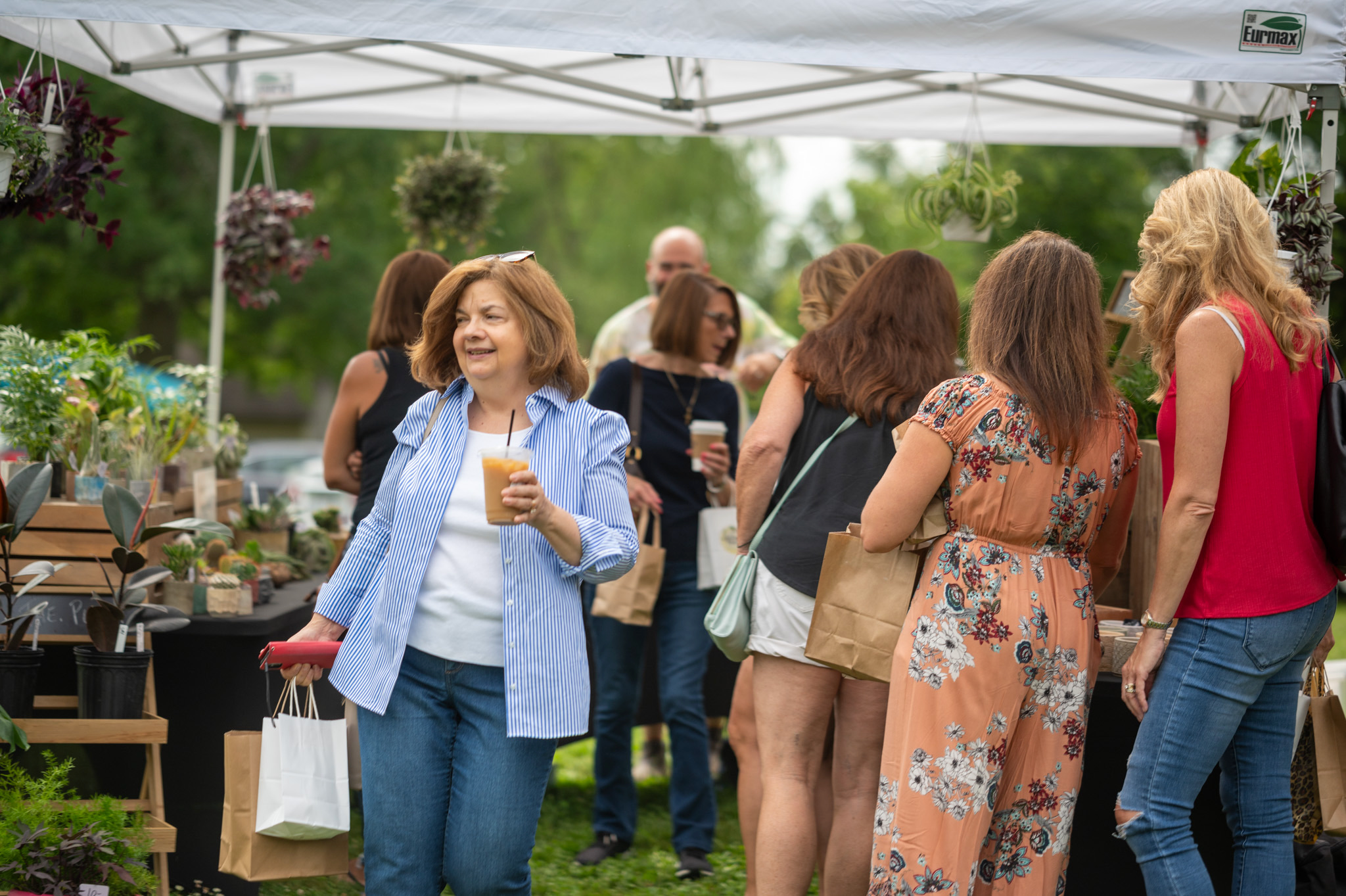 People browsing plants and flowers at an outdoor market under a white canopy tent.