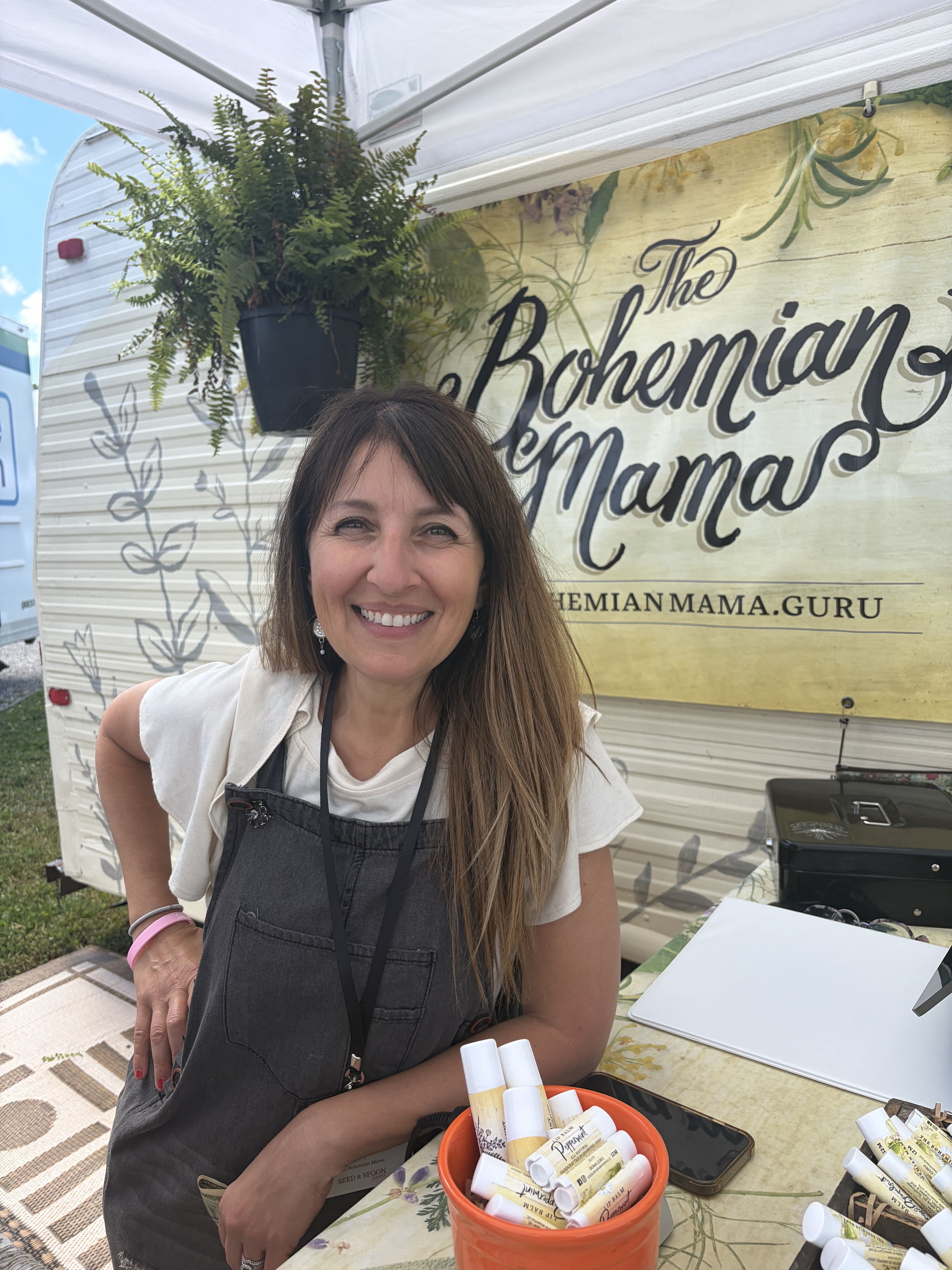 Vivian Brinkman (AKA The Bohemian Mama) smiling at a booth with herbal remedies, surrounded by plants and signs that read 'The Bohemian Mama'.