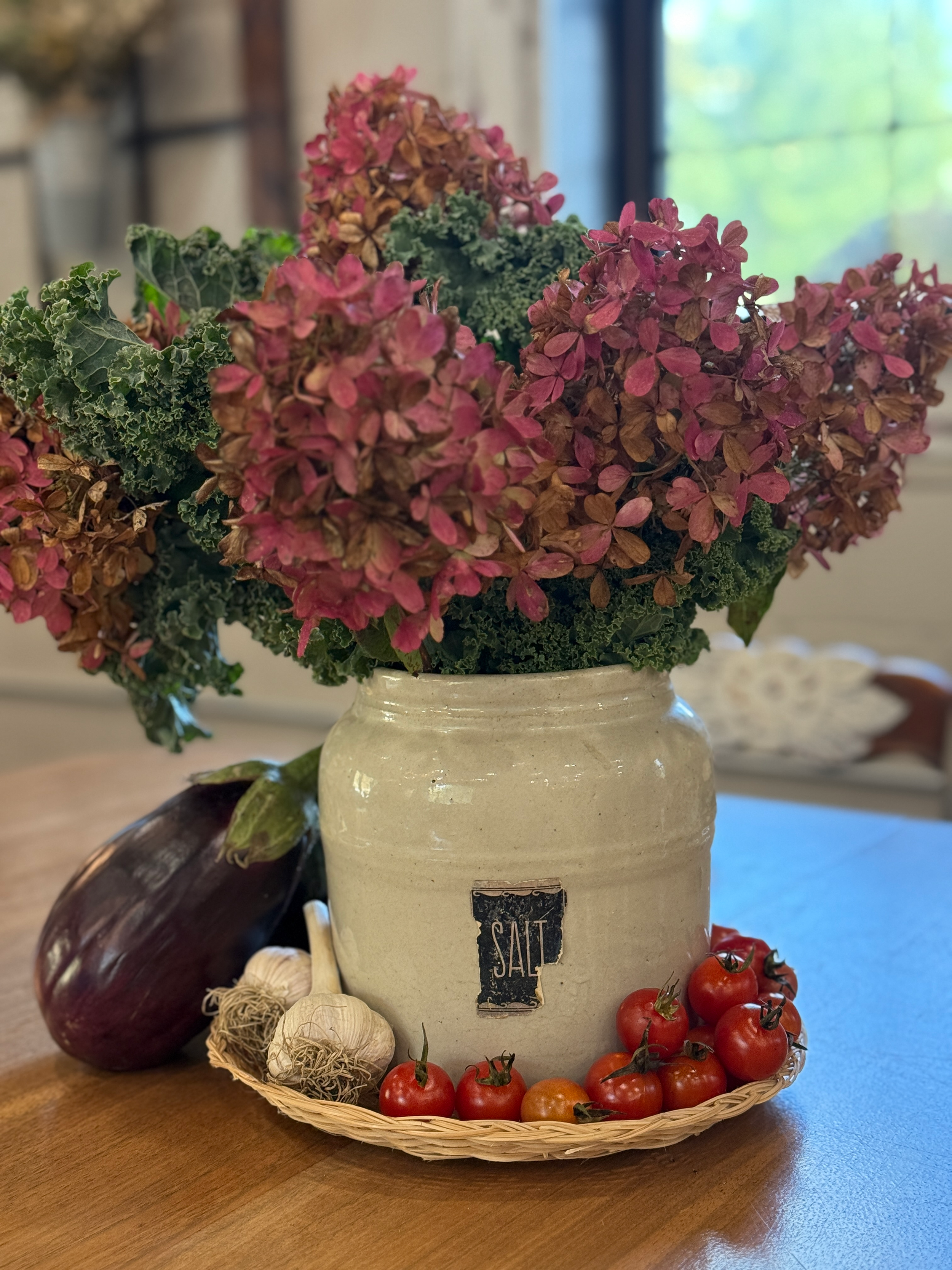 A ceramic salt container with a black label that says 'SALT' sits on a wooden surface. Around it is a wicker basket filled with ripe cherry tomatoes, garlic bulbs, and an eggplant. In the background are blurred kitchen or dining room elements.