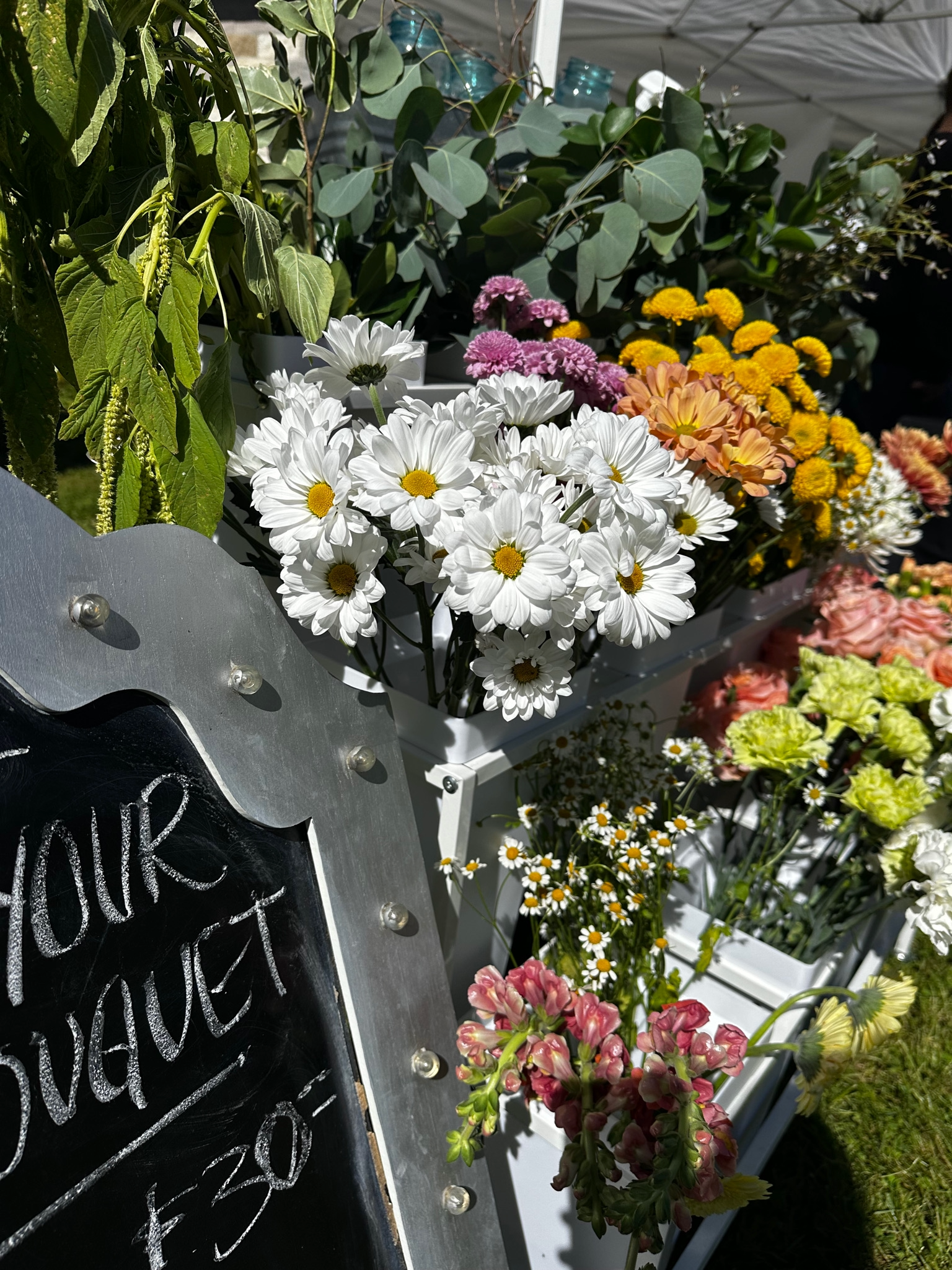 Display of various fresh flowers including white daisies, pink, orange, yellow, and white flowers, with green foliage in the background at an outdoor market stand.