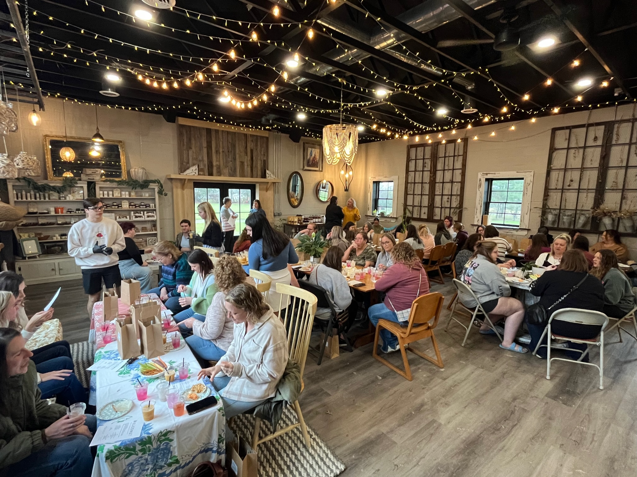 Indoor gathering with people seated at tables, some eating and drinking. The room has string lights on the ceiling, a wooden wall, and large windows. There is a person in white attire near the front, possibly a server, and decorations including framed pictures, mirrors, and hanging light fixtures.