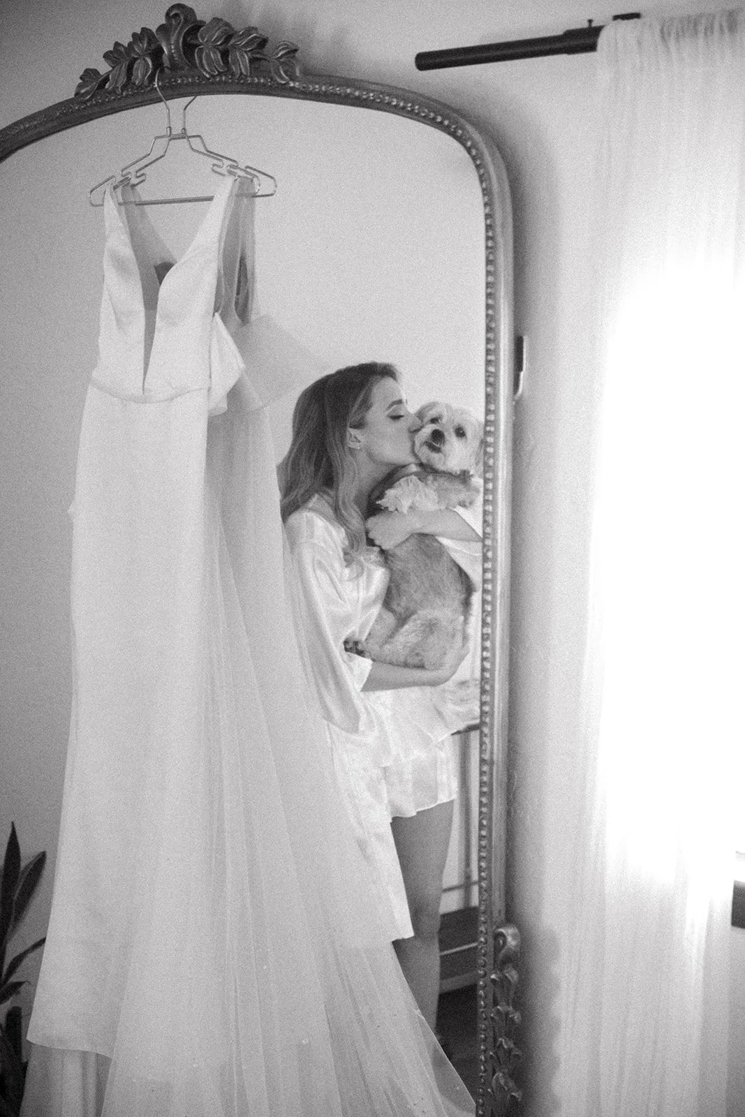 A woman in satin pajamas holding a dog while kissing its face, seen in a mirror reflection, with a wedding dress hanging on the wall.