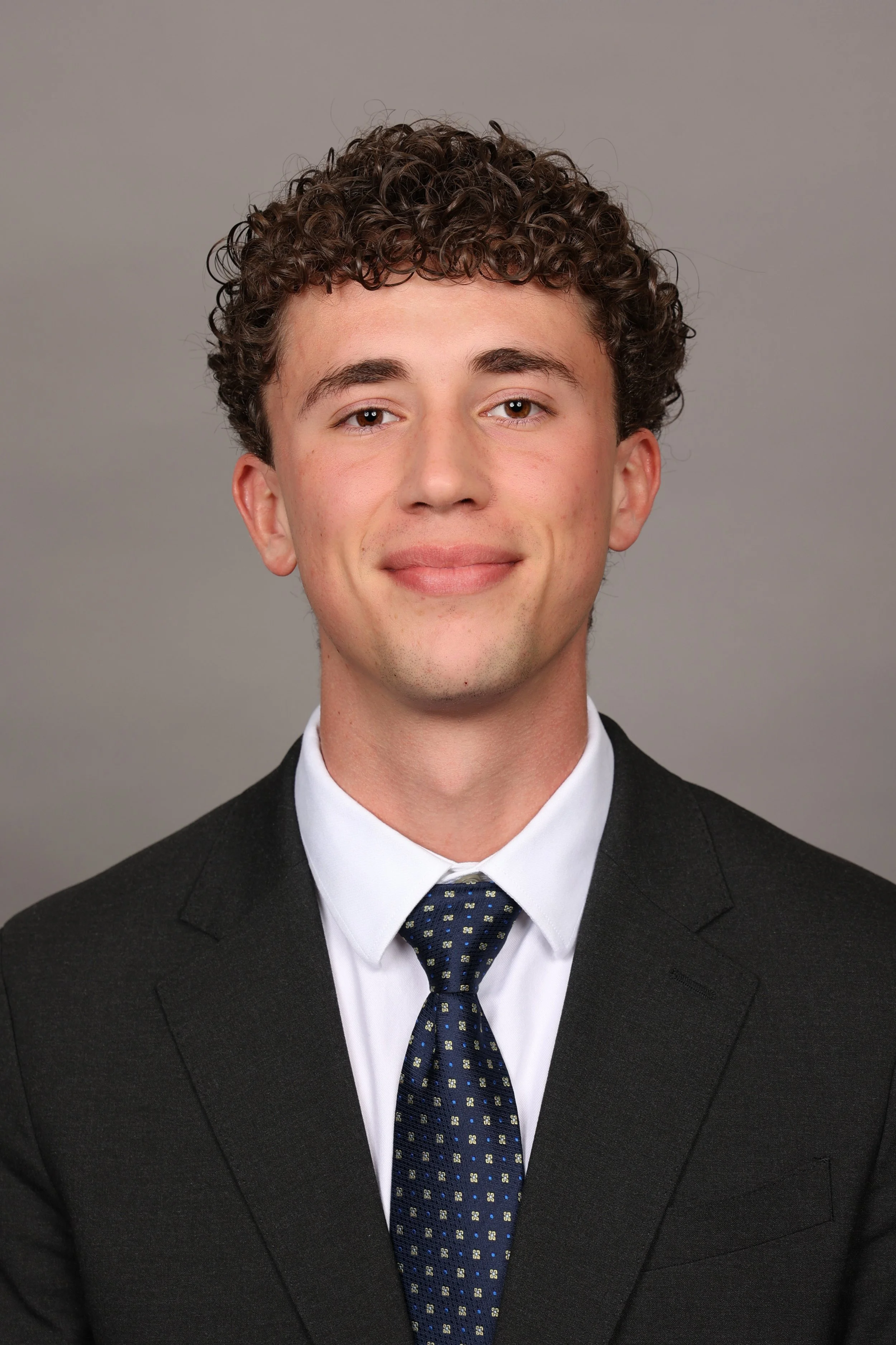 A young man in a business suit smiling outdoors with green grass and trees in the background.