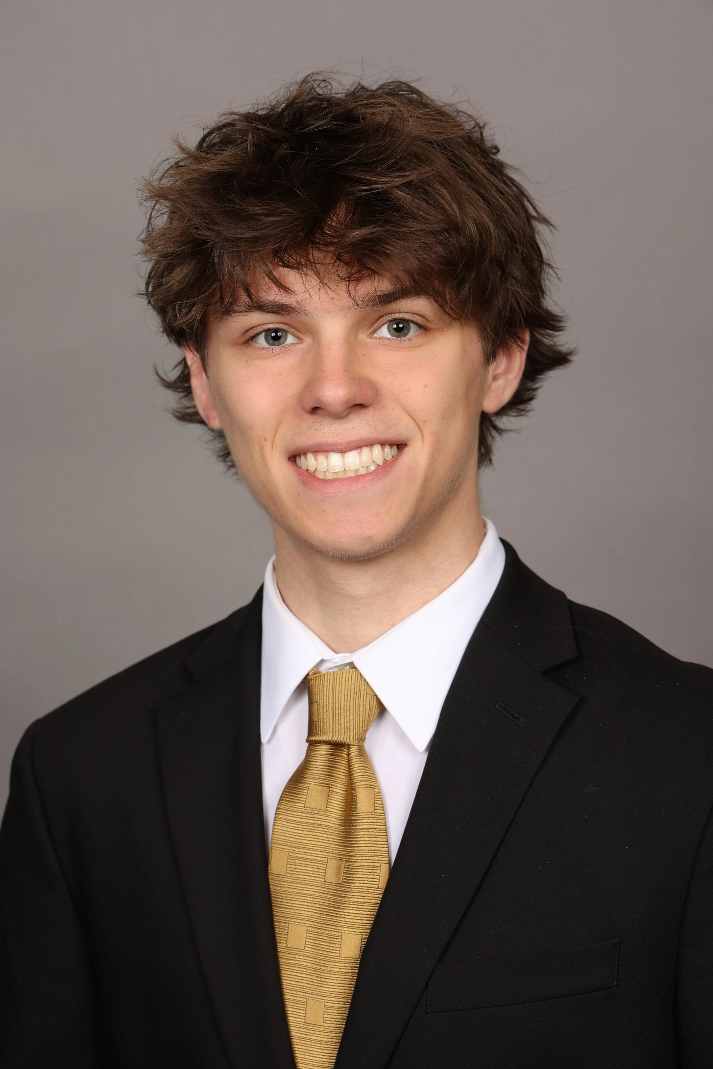 Young woman with long wavy blonde hair, wearing a beige blazer and a gold necklace, smiling against a gray background.