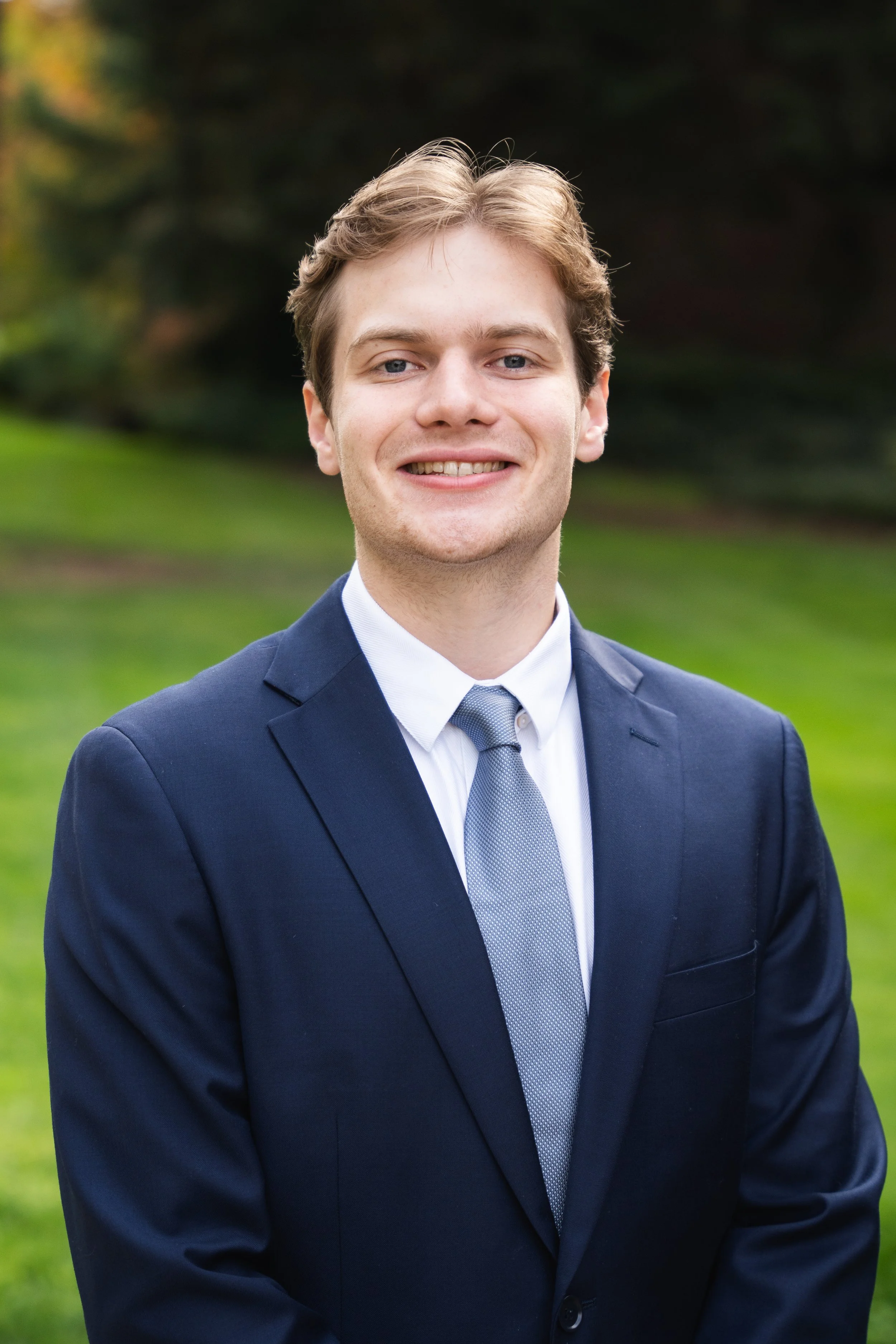 A young man in a light gray suit, white shirt, and black tie, smiling against a neutral background.
