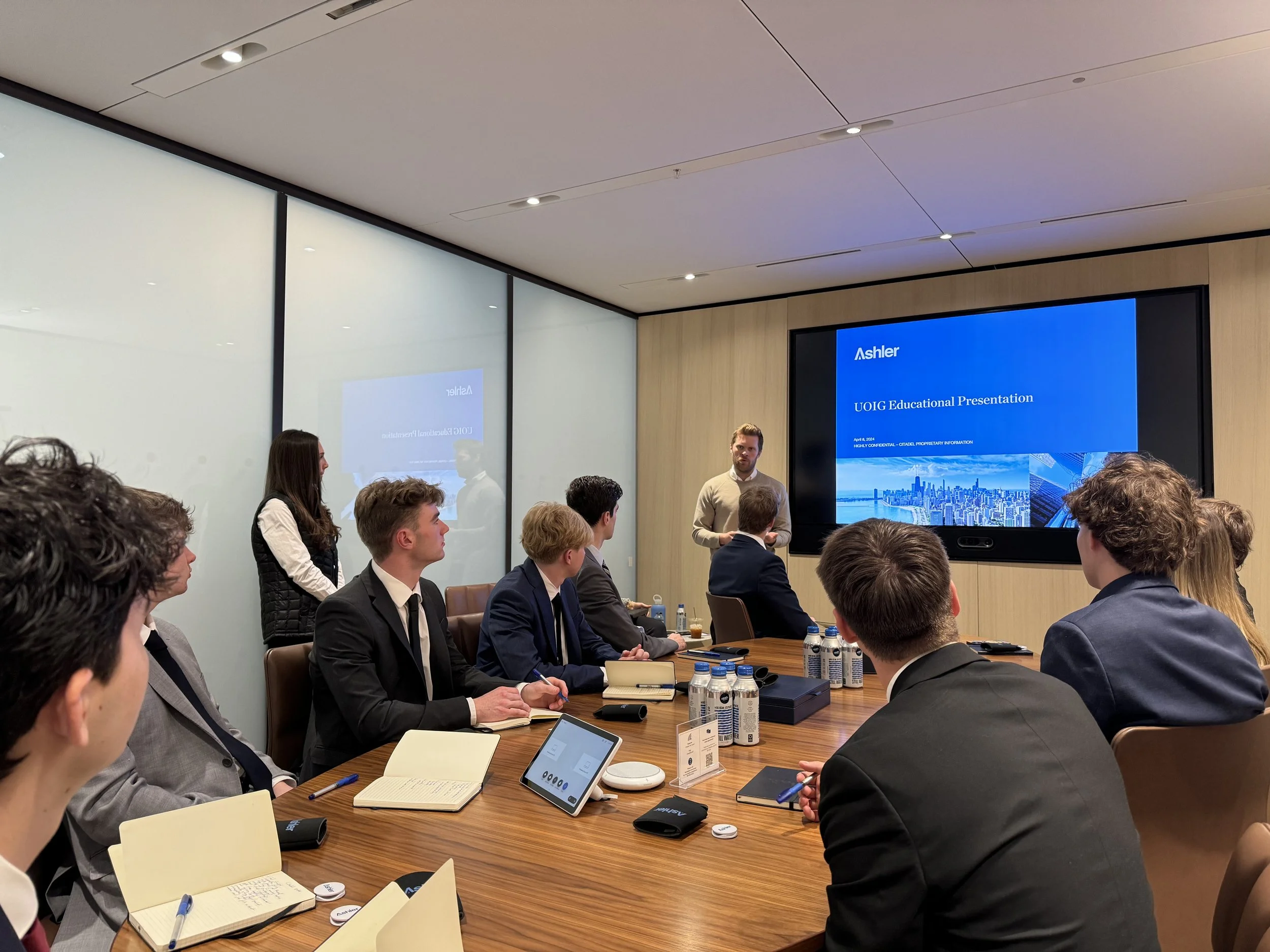 Business meeting in a conference room with a presenter standing near a large screen displaying a presentation titled 'UOIG Educational Presentation'. Attendees are seated around a wooden table, taking notes and listening to the speaker.