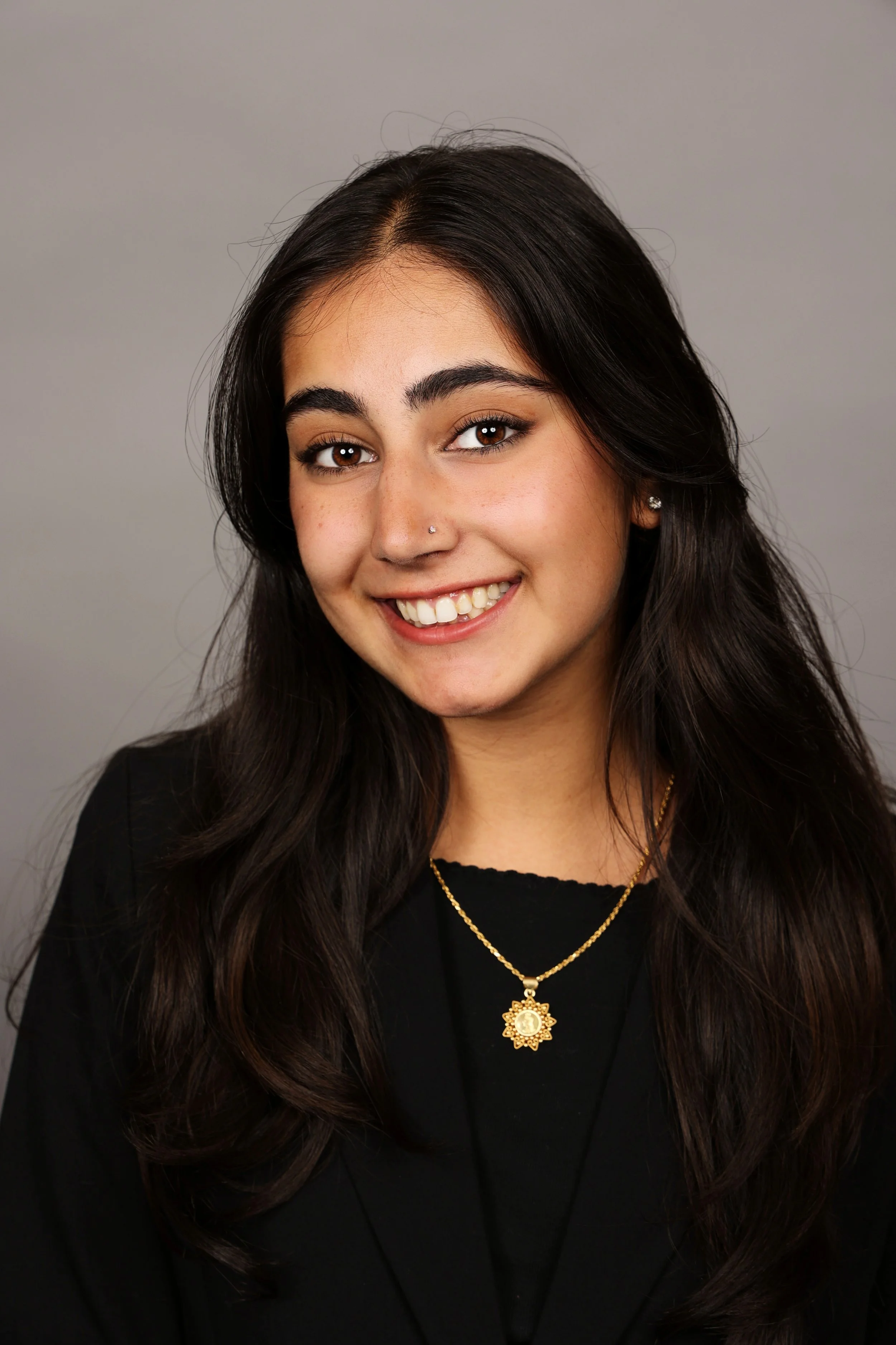 A portrait of a young woman with long dark hair, smiling, wearing earrings, a dark top, and a blazer against a gray background.