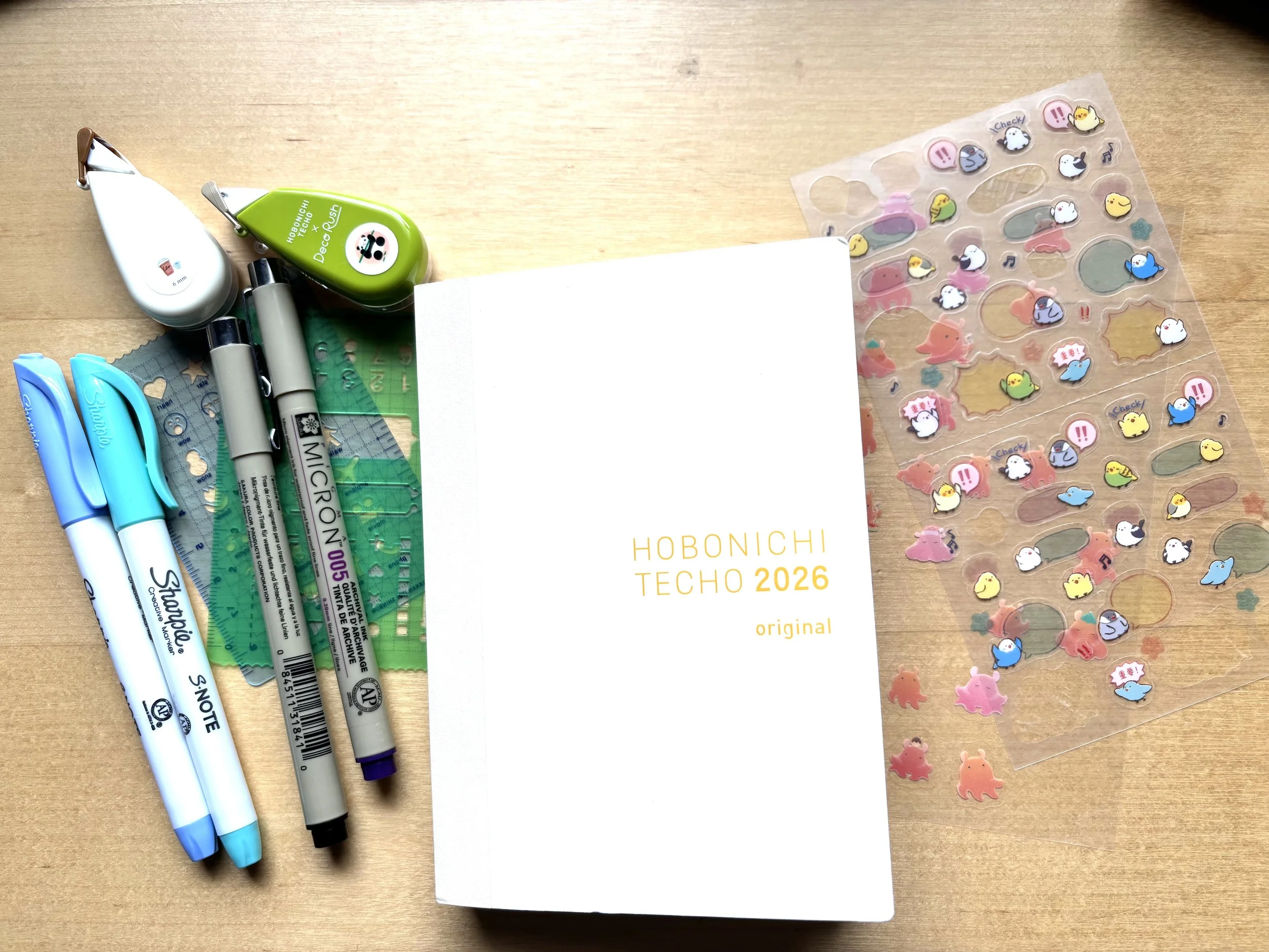 An off-white Hobonichi Techo 2026 original planner in the middle. On the left are two Sharpies in shades of blue, two Micron pens, two stencils, two decorate tapes. On the right are two sheets of clear stickers with octopuses and birds.