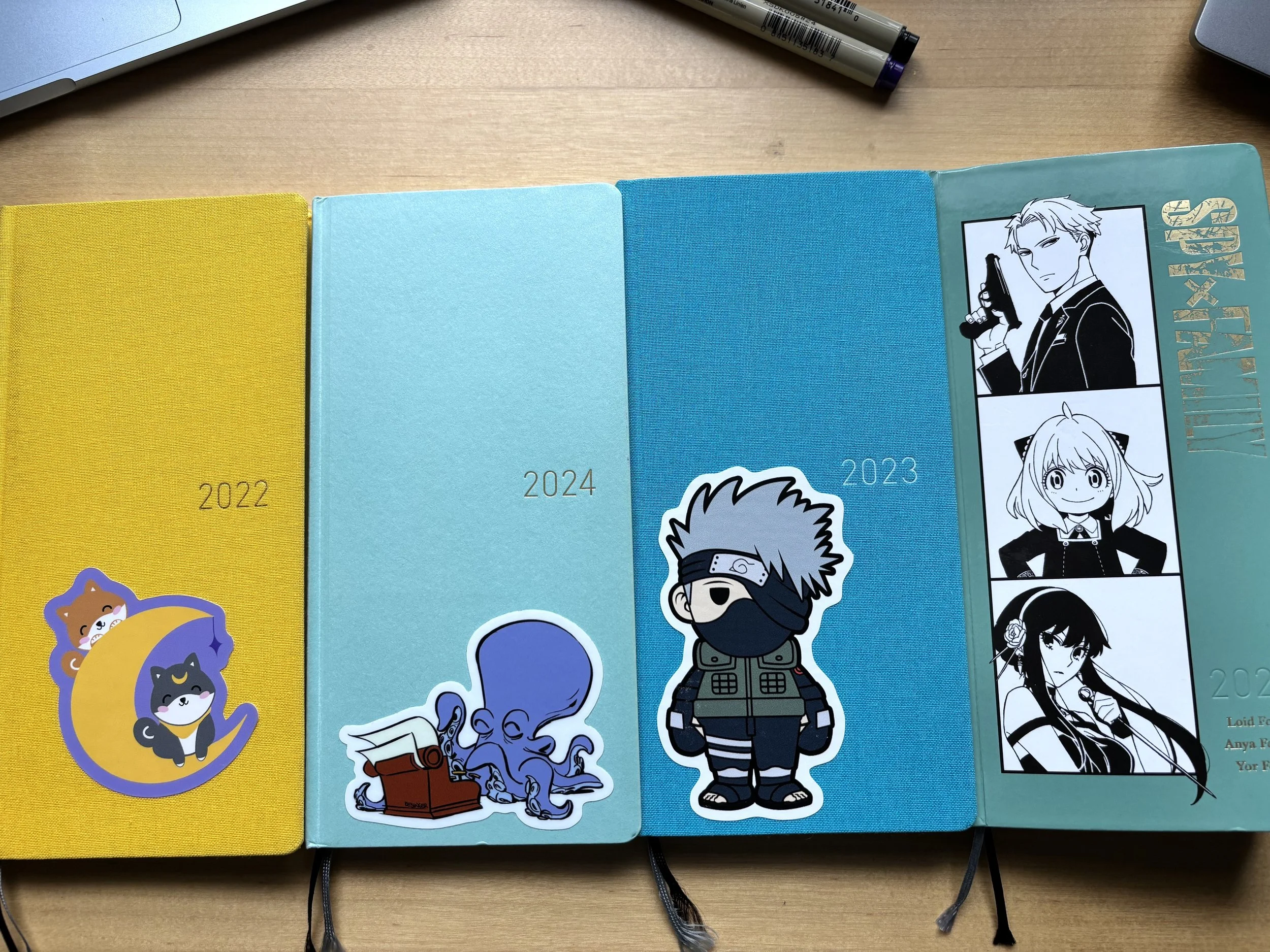 Four Hobonichi Techo Weeks with a cute sticker on each. From left to right, 2022 in yellow, 2024 in aqua, 2023 in blue, and 2025 in teal with a Spy x Family design.