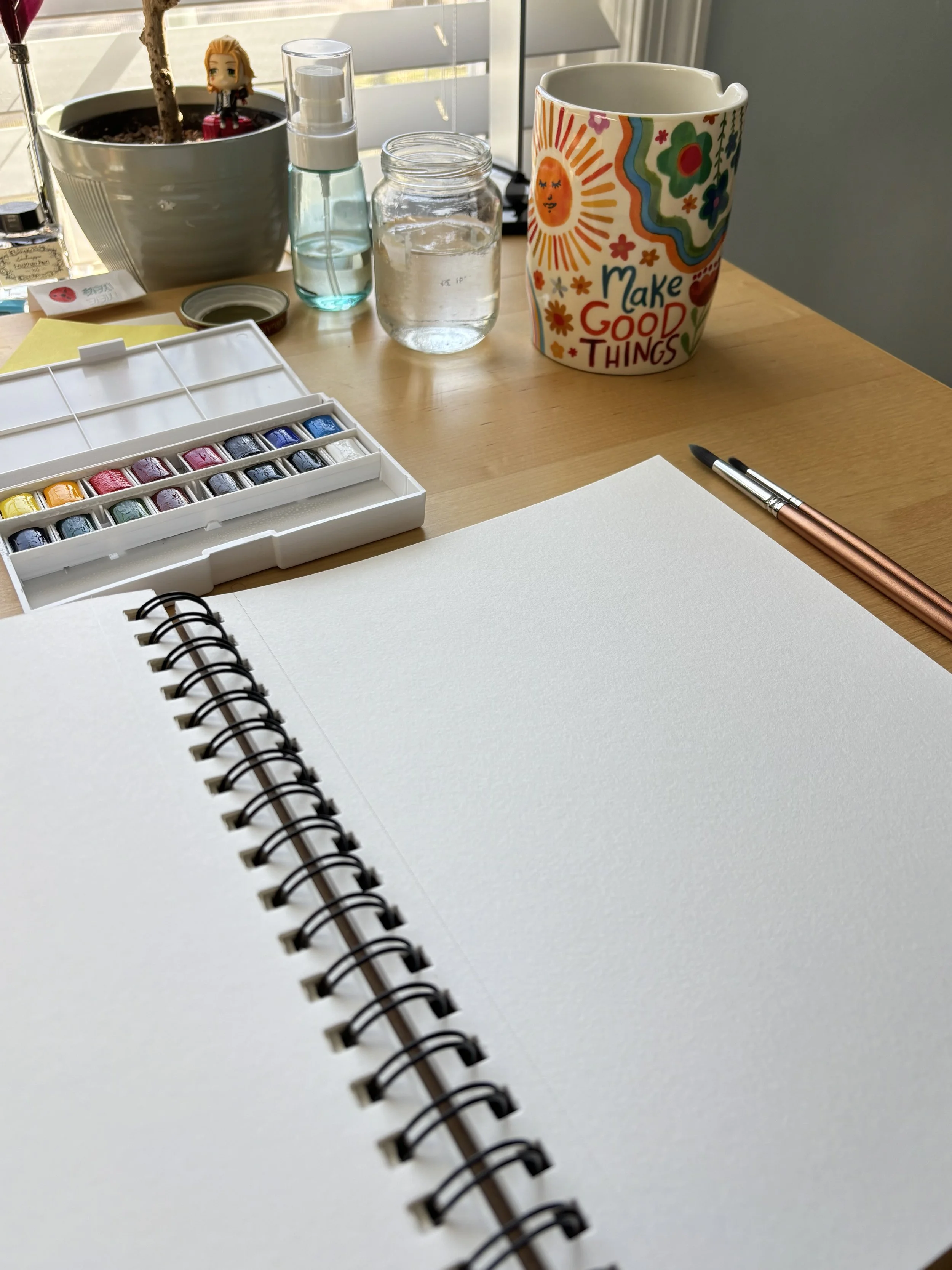 A picture of a desk where an open sketchbook is laid out. There is an open pan of watercolor paints in a rainbow of colors, some paint brushes, a jar of clear water, and a ceramic cup with a sunshine and flowers on it and says "Make Good Things."