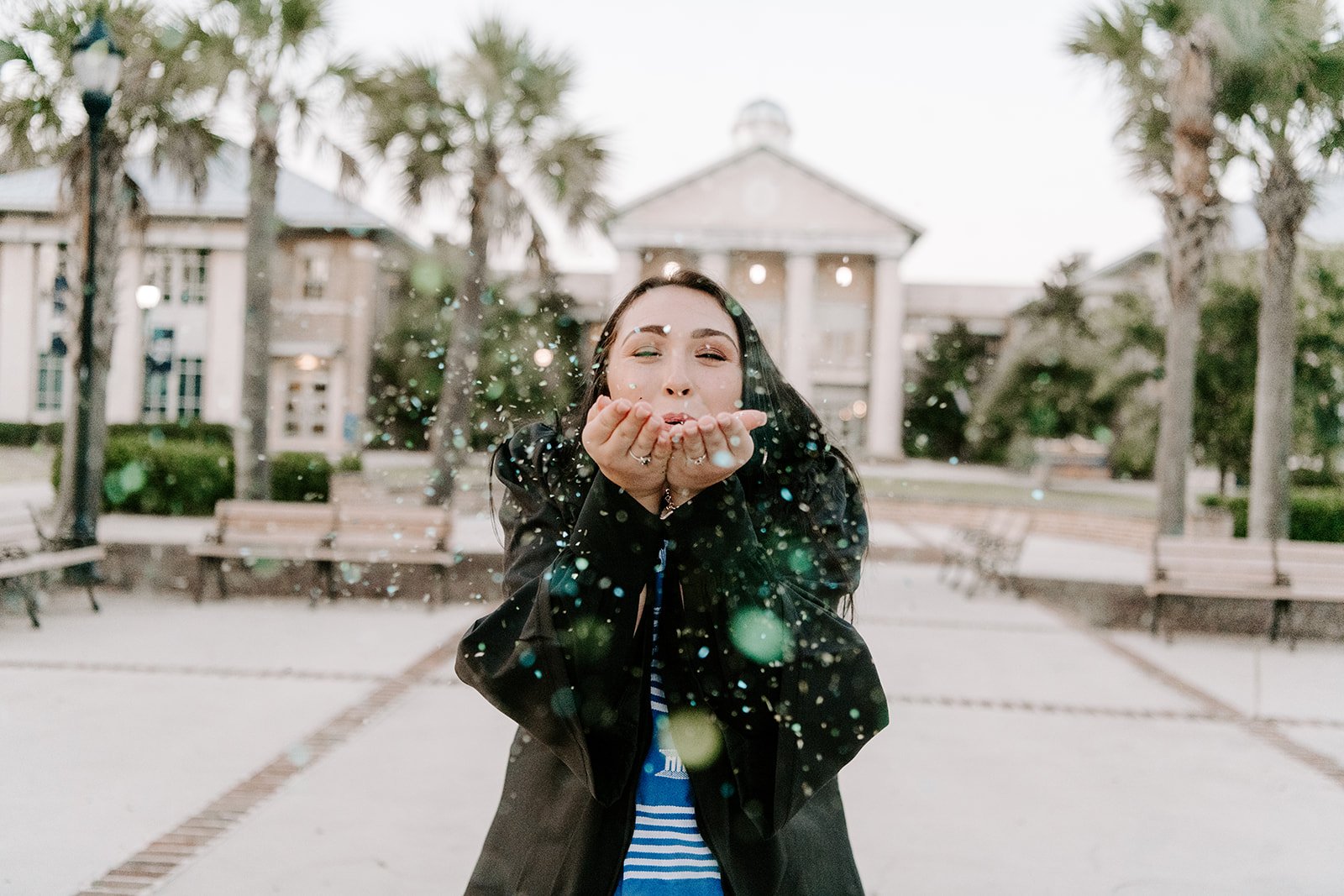 A graduate in a black gown blowing green and blue glitter at USC Beaufort’s Hilton Head Gateway Campus in Bluffton, SC. The background shows the iconic domed building and Lowcountry palmetto trees.