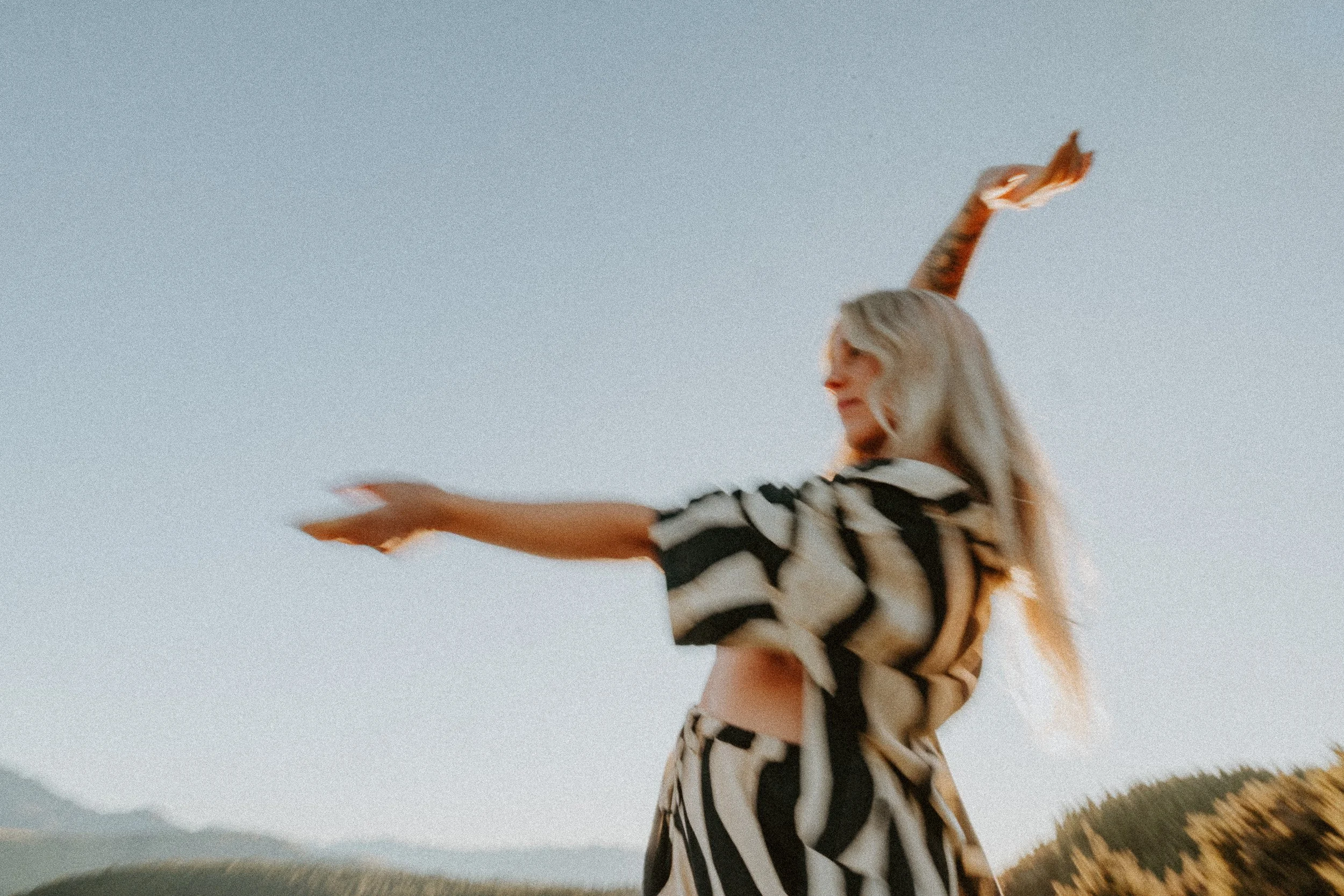 A woman with long blonde hair, wearing a zebra print outfit, is standing outdoors with mountains and a clear sky in the background. She is smiling and extending her arms outward.