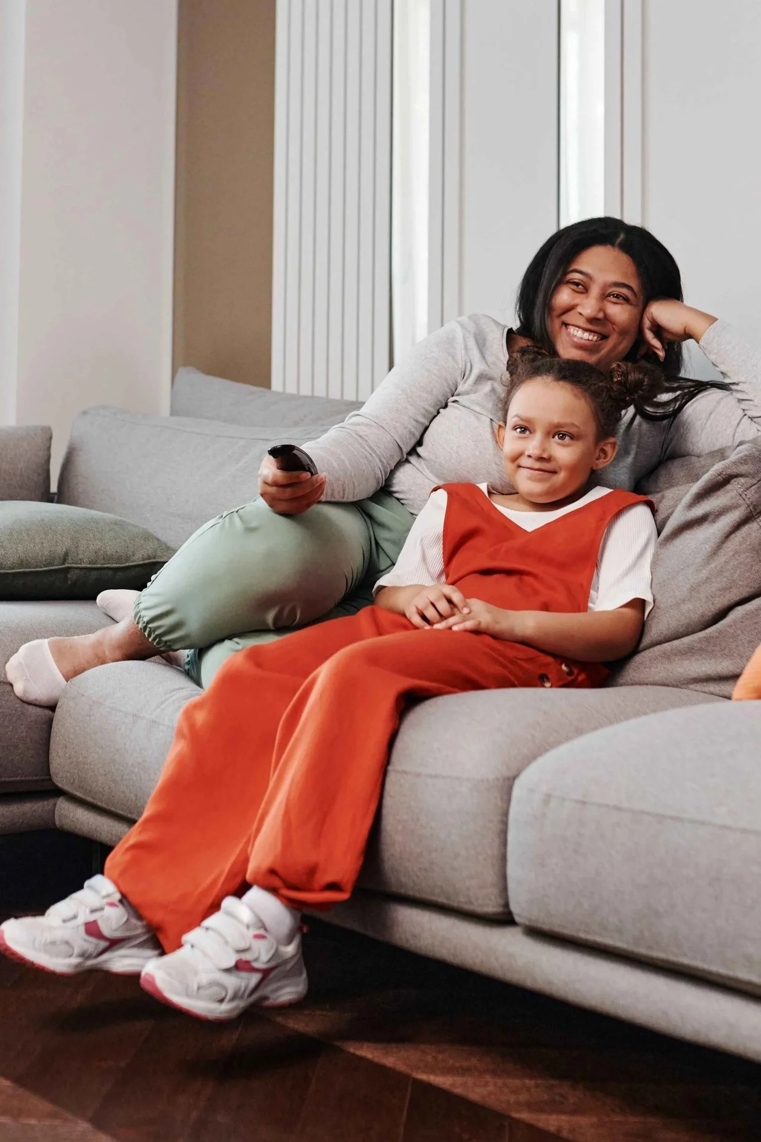 Woman and young girl smiling on a gray couch while watching TV together, comfort shows Hoboken NJ counselor