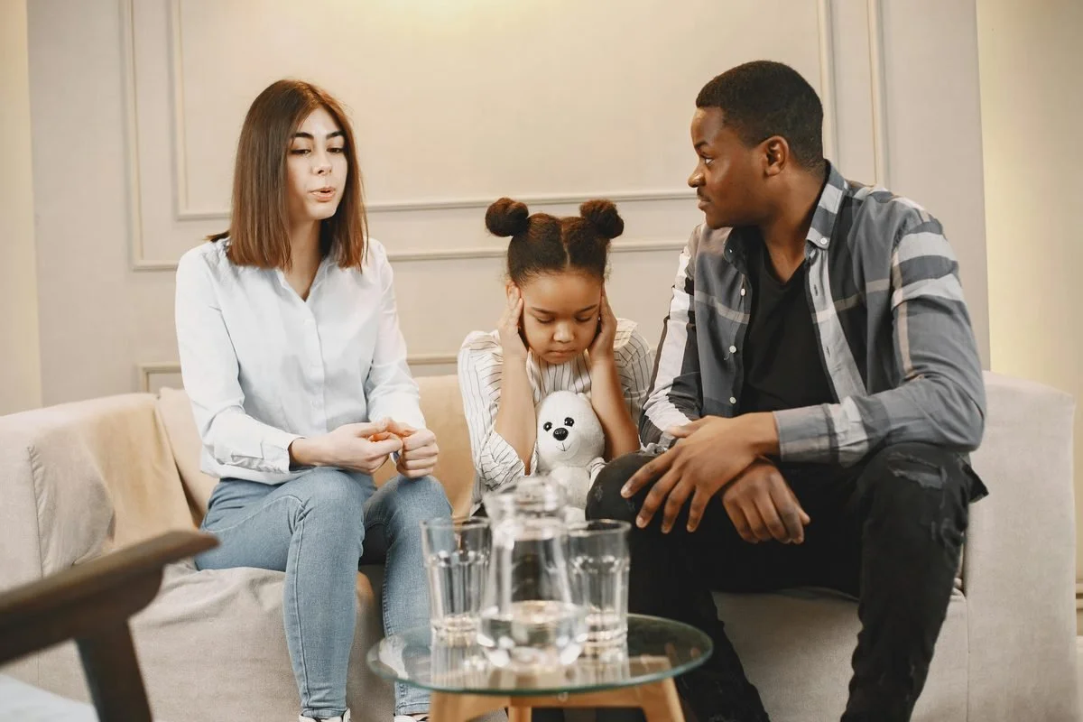 A young girl covers her ears looking distressed while sitting between her parents during a family therapy session, representing the emotional impact of conditional love in a dysfunctional family