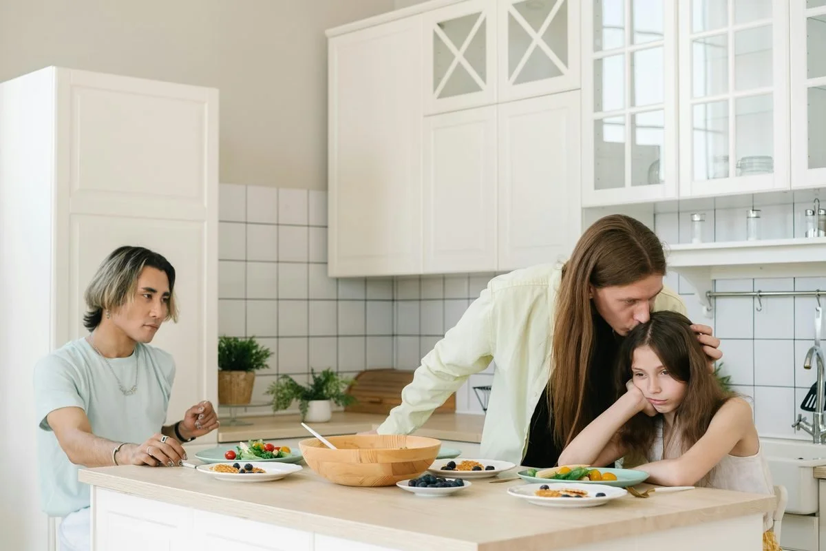 A parent leans over to comfort a visibly upset child at the kitchen counter during mealtime while another family member looks on, illustrating emotional neglect and conditional love in a dysfunctional family system