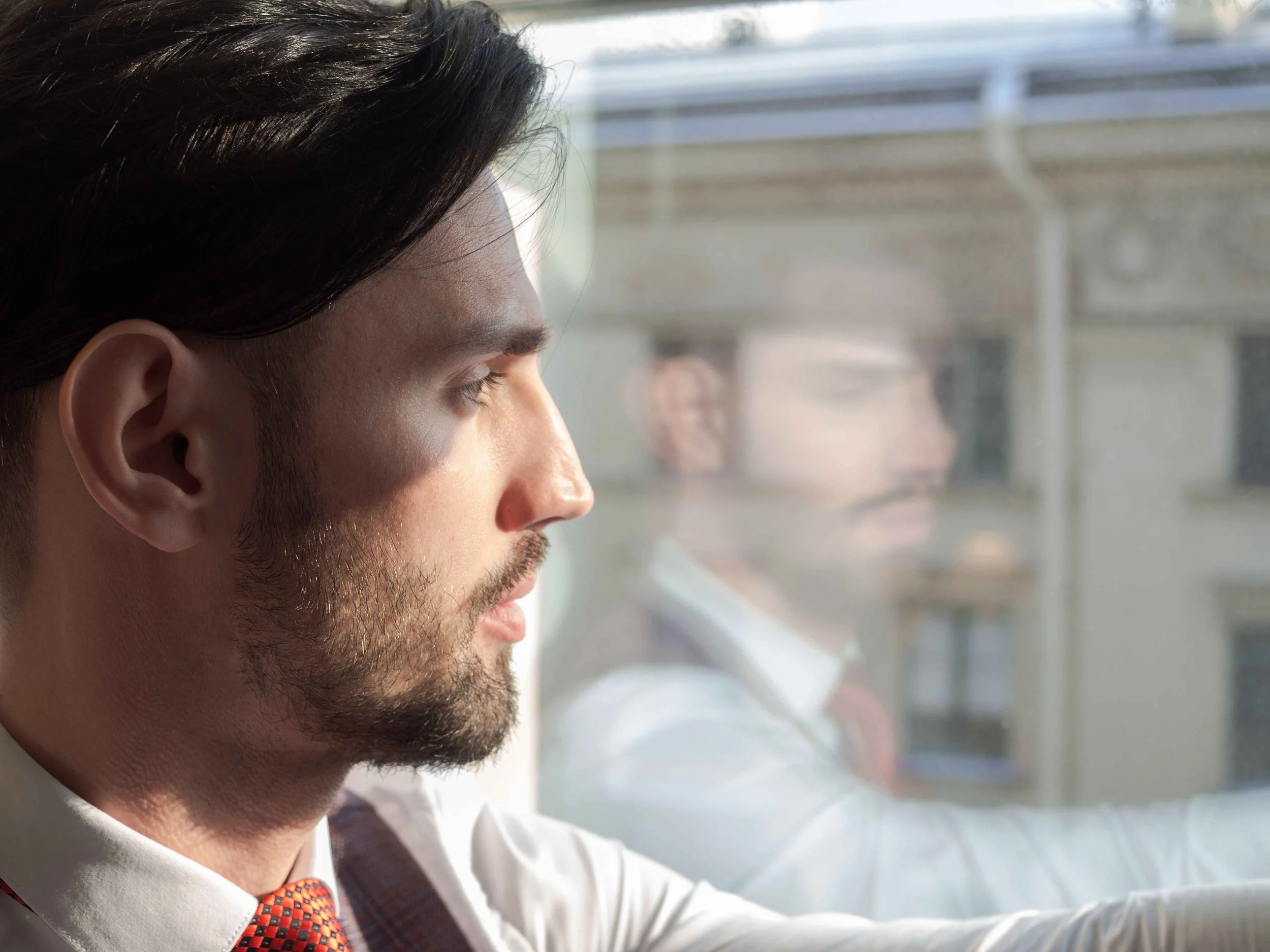 Man looking thoughtfully out a window with his reflection in the glass, the kind of self-reflection that separates narcissistic traits from NPD, Hoboken NJ therapist
