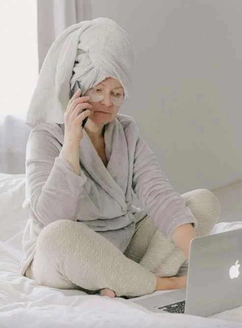 older woman doing under eye mask and on the phone with towel and robe on bed while bed rotting in jersey city nj