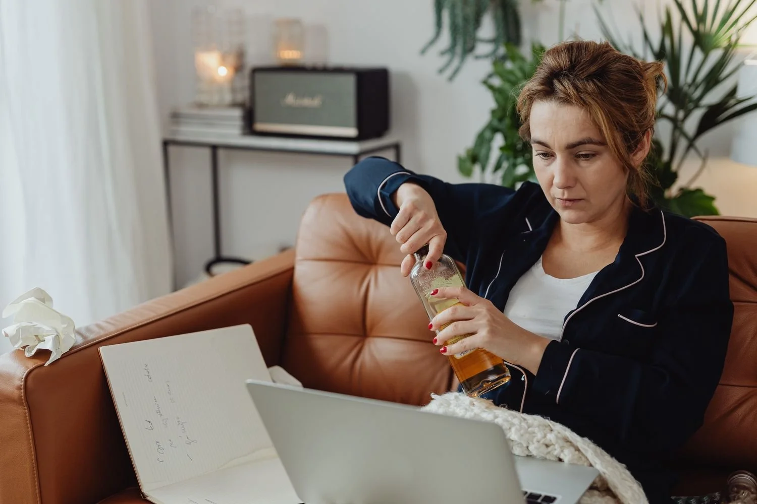 Woman in pajamas opening a bottle of liquor on a leather couch while working on her laptop, high functioning alcoholic Hoboken NJ therapist