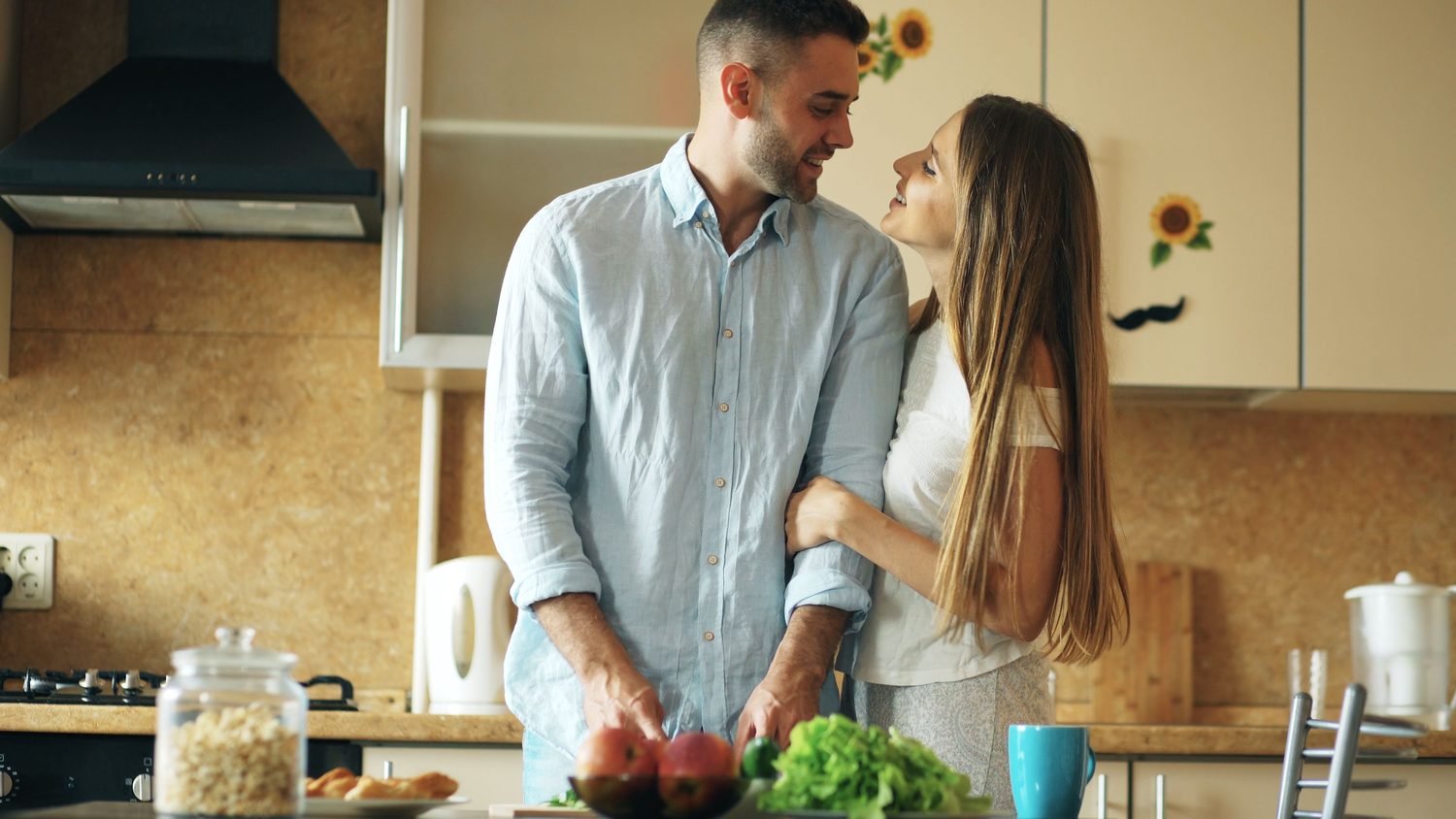 Woman smiling up at her partner while he chops vegetables in a sunny kitchen, why healthy relationships feel boring Hoboken NJ counselor