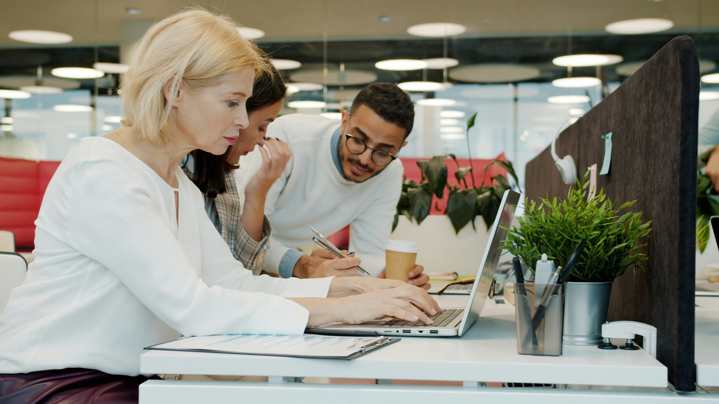 Mixed-age office team reviewing work together at a laptop in an open-plan workspace.