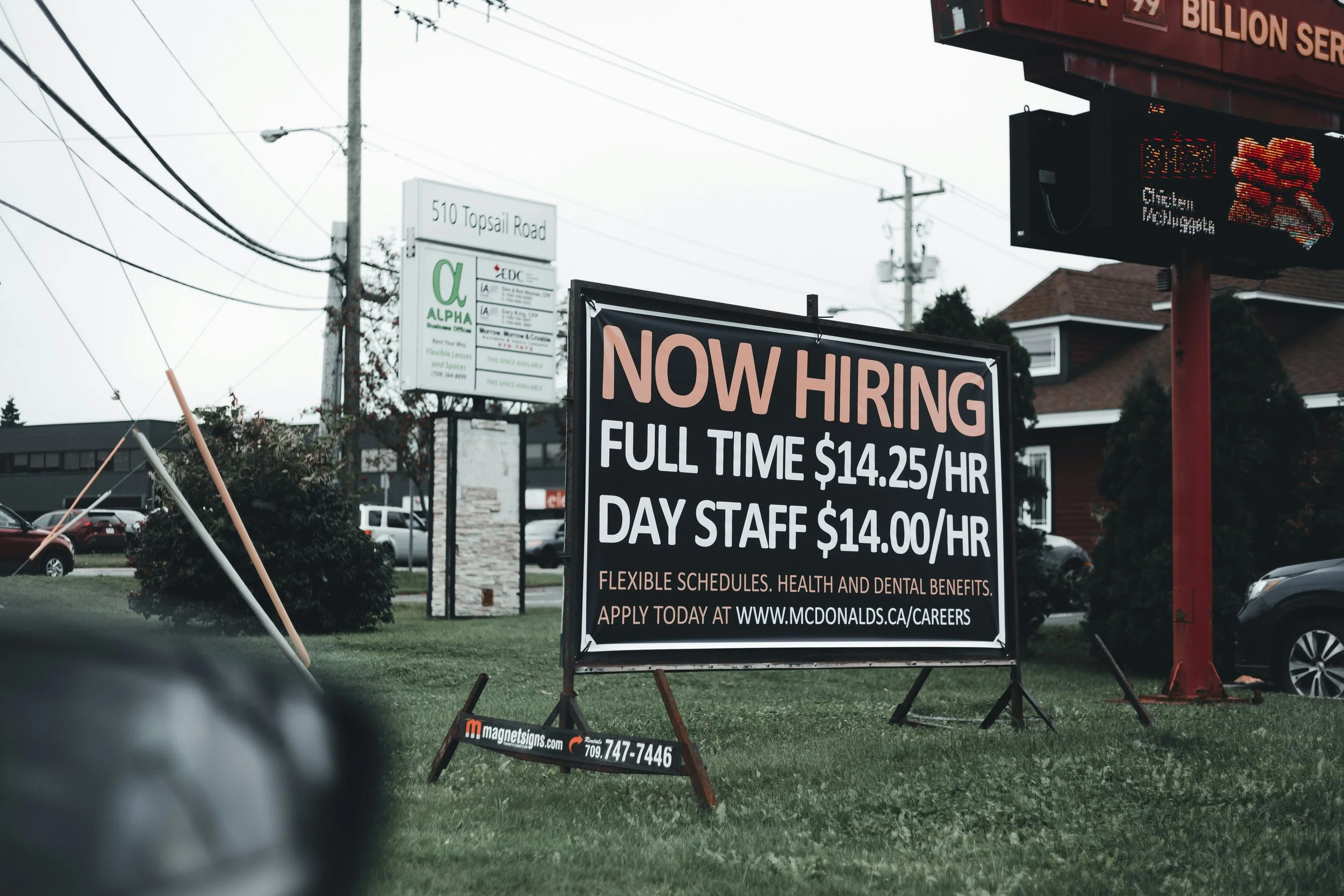 Roadside “Now Hiring” sign advertising low-wage hourly jobs outside a fast-food restaurant.