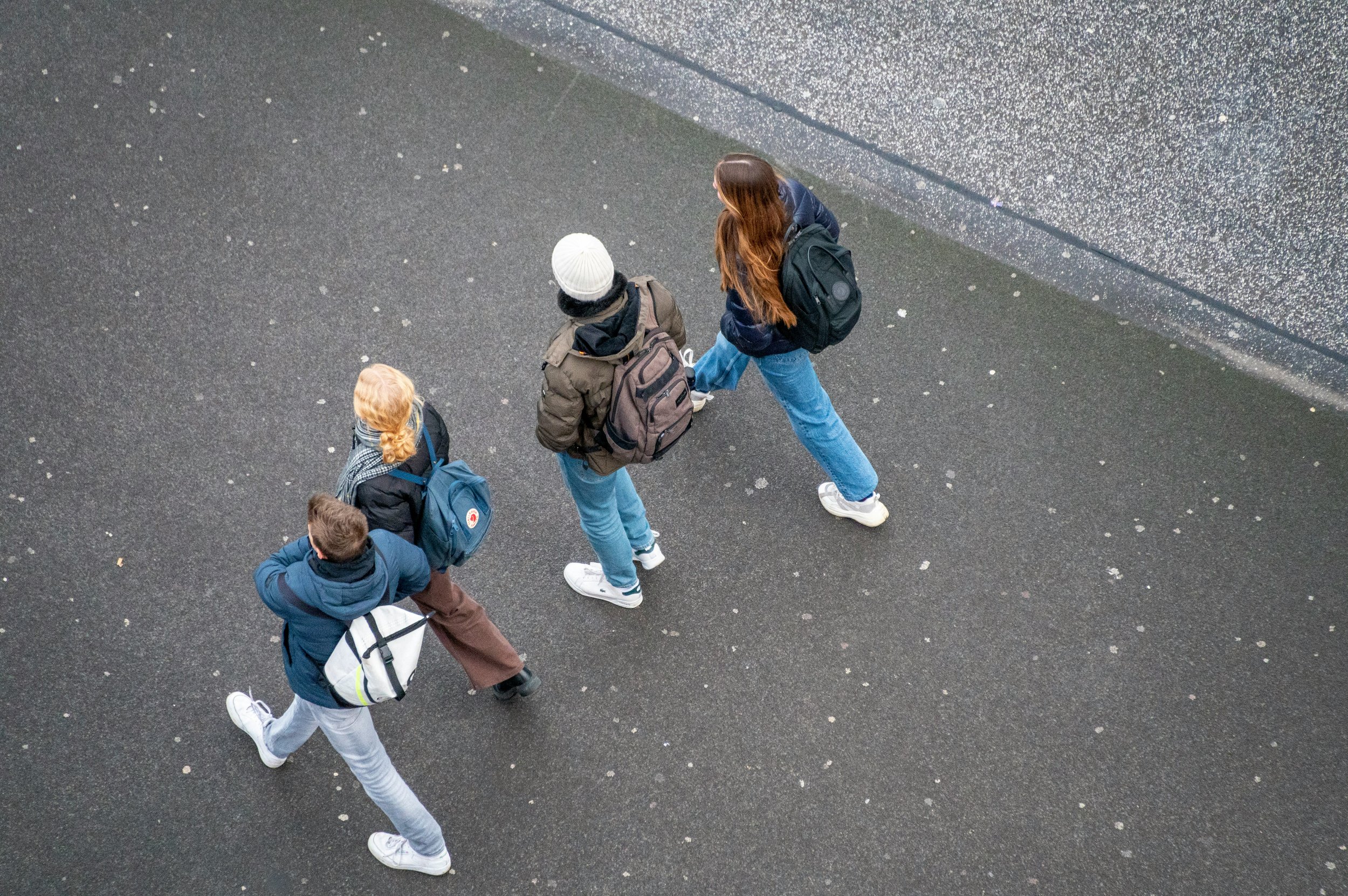 Young people walking outdoors on a grey pavement, seen from above, each carrying a backpack.
