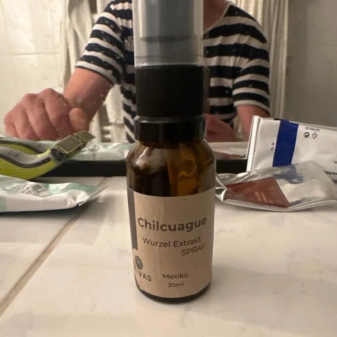 A small bottle labeled 'Chilcuague Wurzel Extrakt SPRAY' with a spray nozzle on top, sitting on a table with various items around it. A person in a striped shirt is in the background, slightly out of focus.