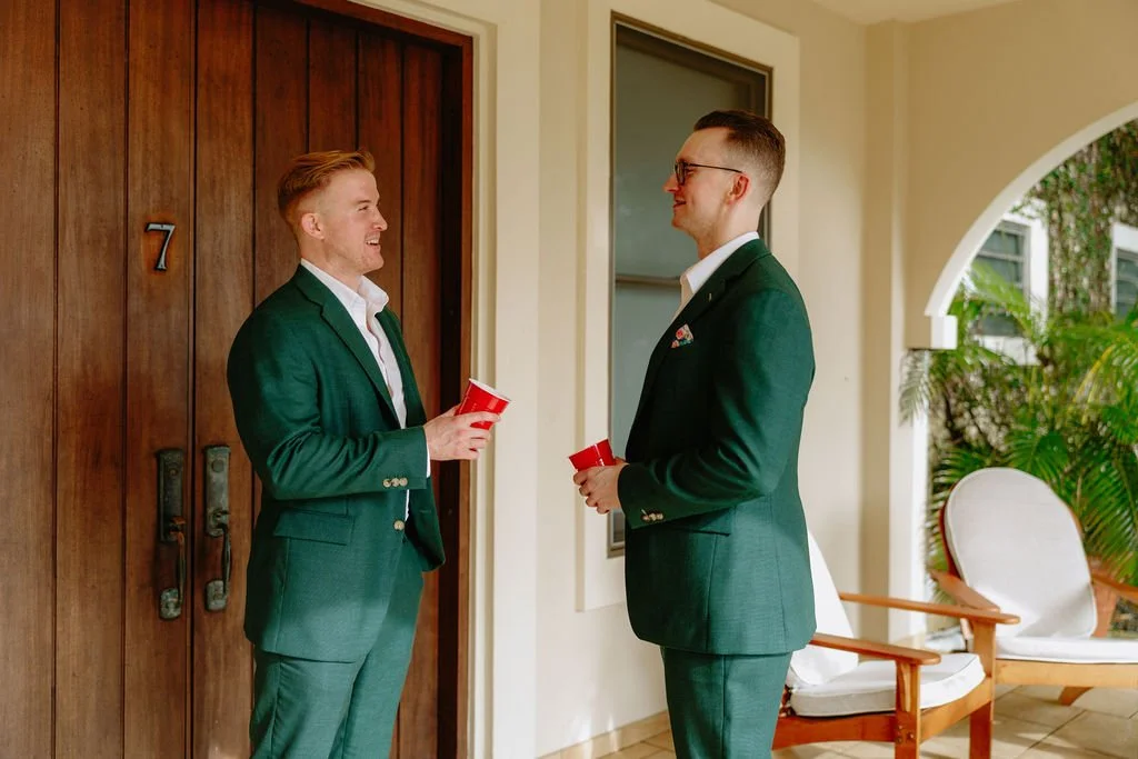 Two men in green suits standing and talking outside a house, holding red cups, near a wooden door and a white outdoor chair.