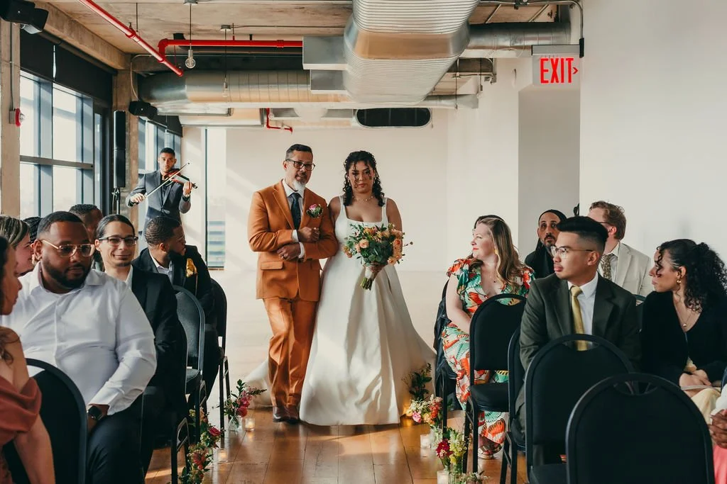 A wedding ceremony with a bride and groom walking down the aisle, surrounded by seated guests and a musician playing violin in a modern, industrial-style venue.