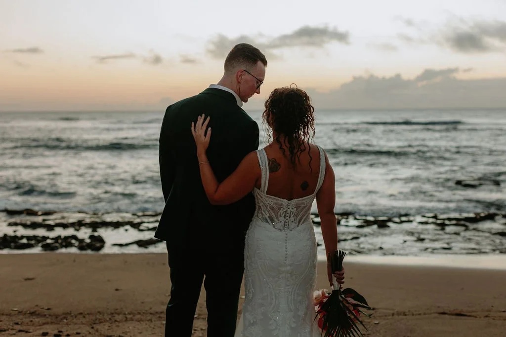 A couple dressed in wedding attire standing on a beach during sunset, with the ocean in the background. The bride is holding a bouquet and has tattoos on her back, while the groom is wearing a suit and glasses.