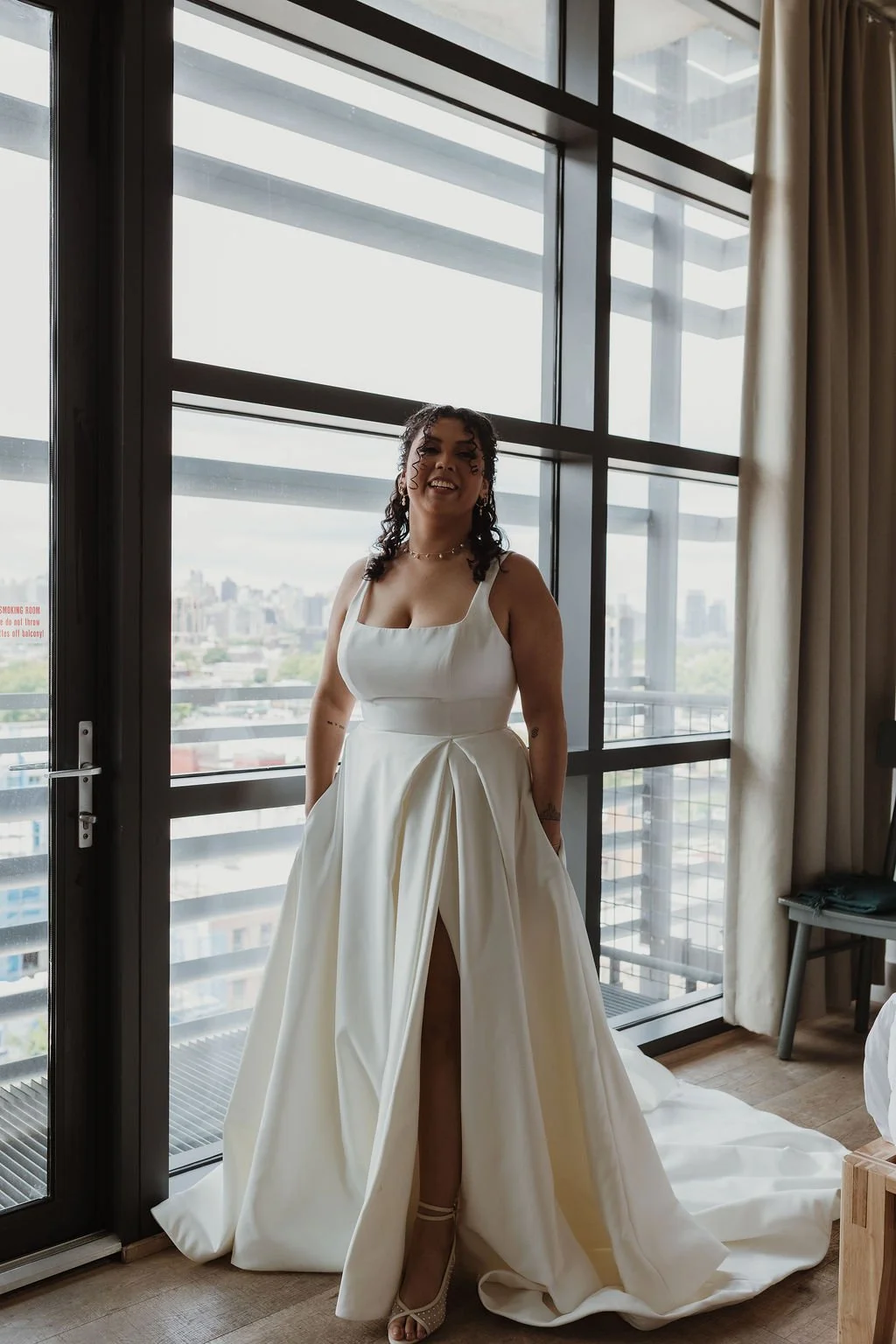 Woman in a wedding dress standing inside near a large window with a city view outside.