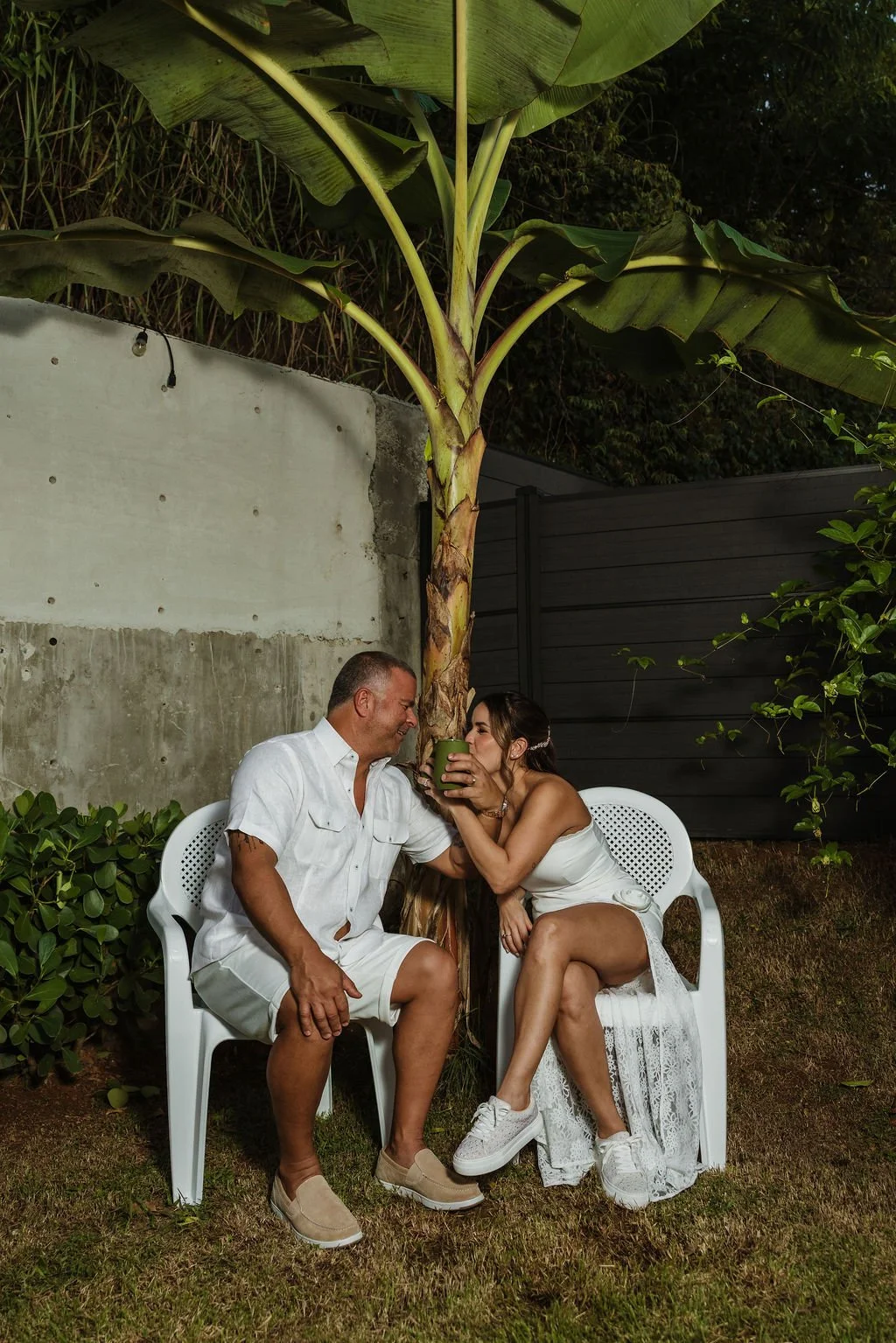 A couple sitting on white plastic chairs outside, sharing a drink under a large banana plant, with greenery and a concrete wall in the background.