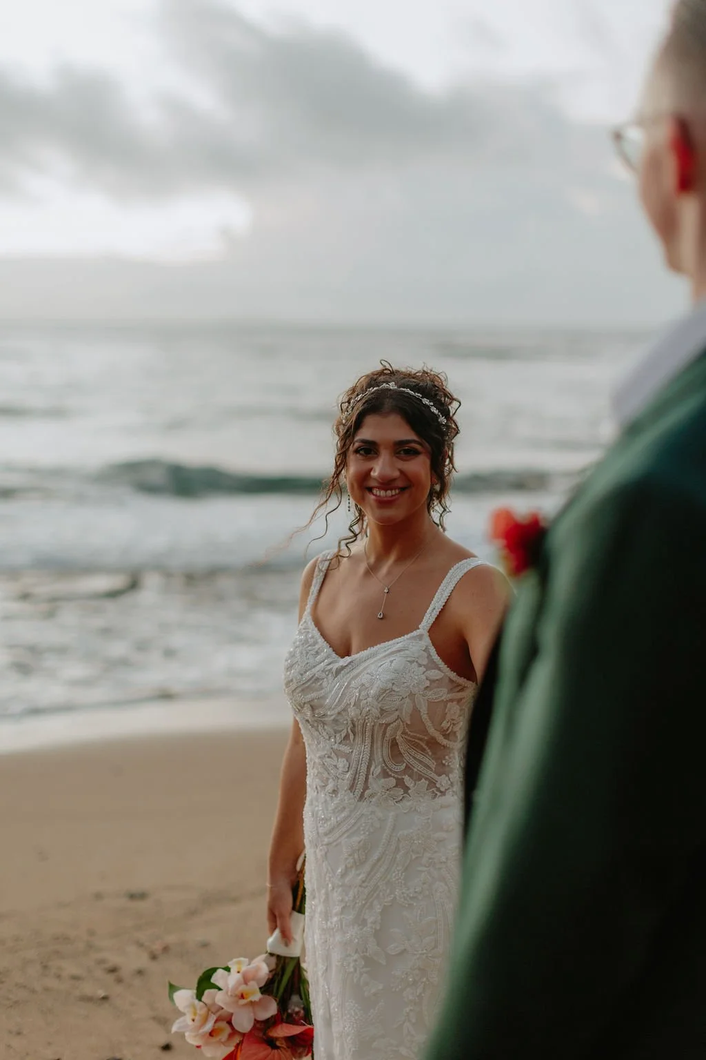 Smiling bride in a lace wedding dress holding a bouquet of pink and white flowers on the beach during cloudy weather, with a person partially visible in the foreground.