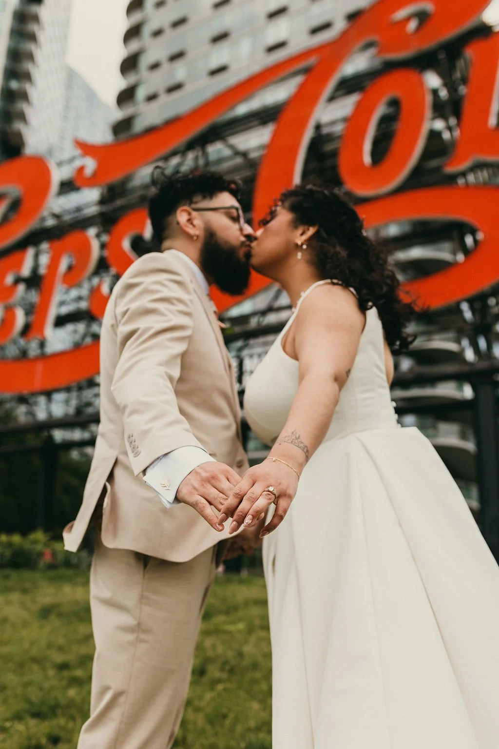 A couple dressed in wedding attire kissing outdoors in front of a large red sign that says "LOVE" with city buildings in the background.