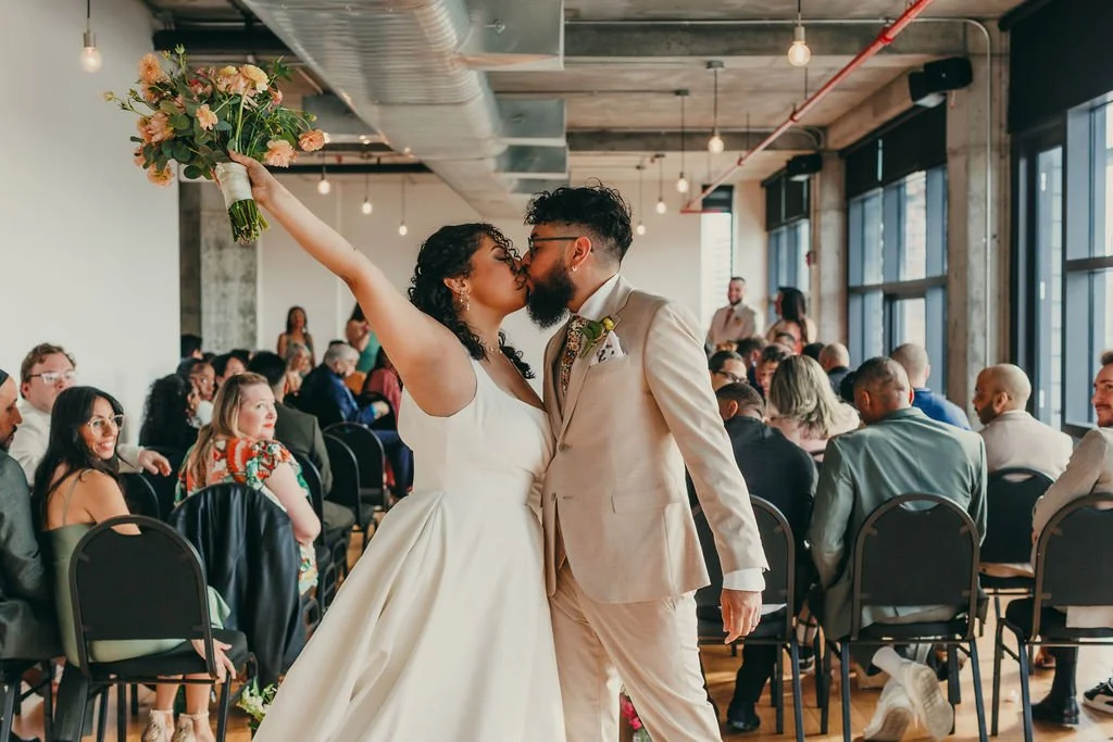 A bride and groom kissing at their wedding ceremony, with the bride holding a bouquet of flowers raised in the air. Guests are seated in the background in a modern, well-lit venue.