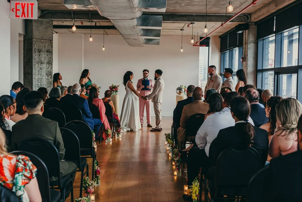 Couple exchanging vows during a wedding ceremony in an industrial-style venue with guests seated on both sides and large windows on the right.