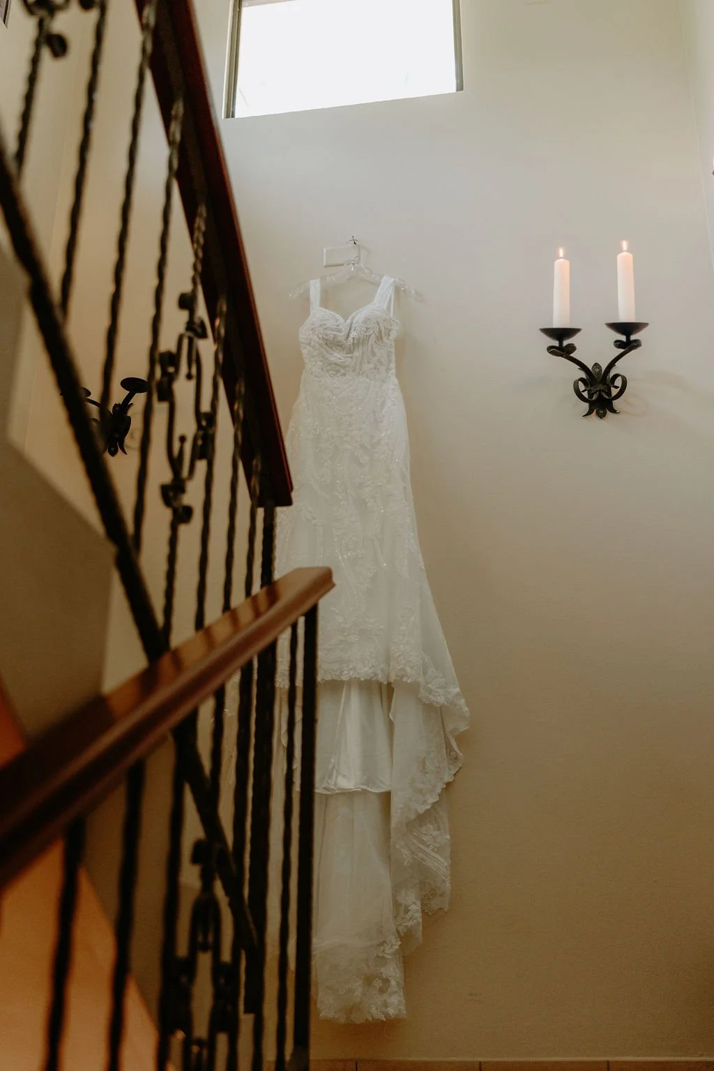 A white lace wedding dress hanging on a clear hanger on a wall, facing a staircase with a black iron railing and a wooden handrail, and a wall-mounted black candle holder with two lit white candles.