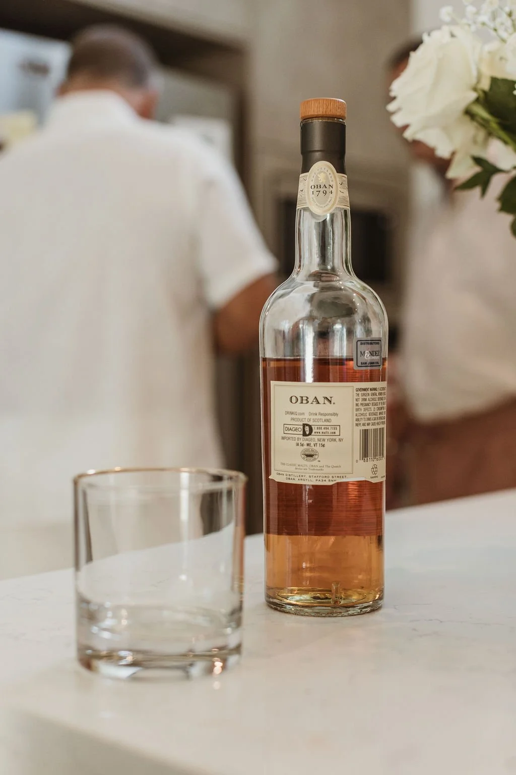 A bottle of rosé wine labeled "Oban" standing on a white surface with a clear glass in front of it. In the blurred background, there are two people, one in a white shirt and the other partially visible, near a bouquet of white flowers.